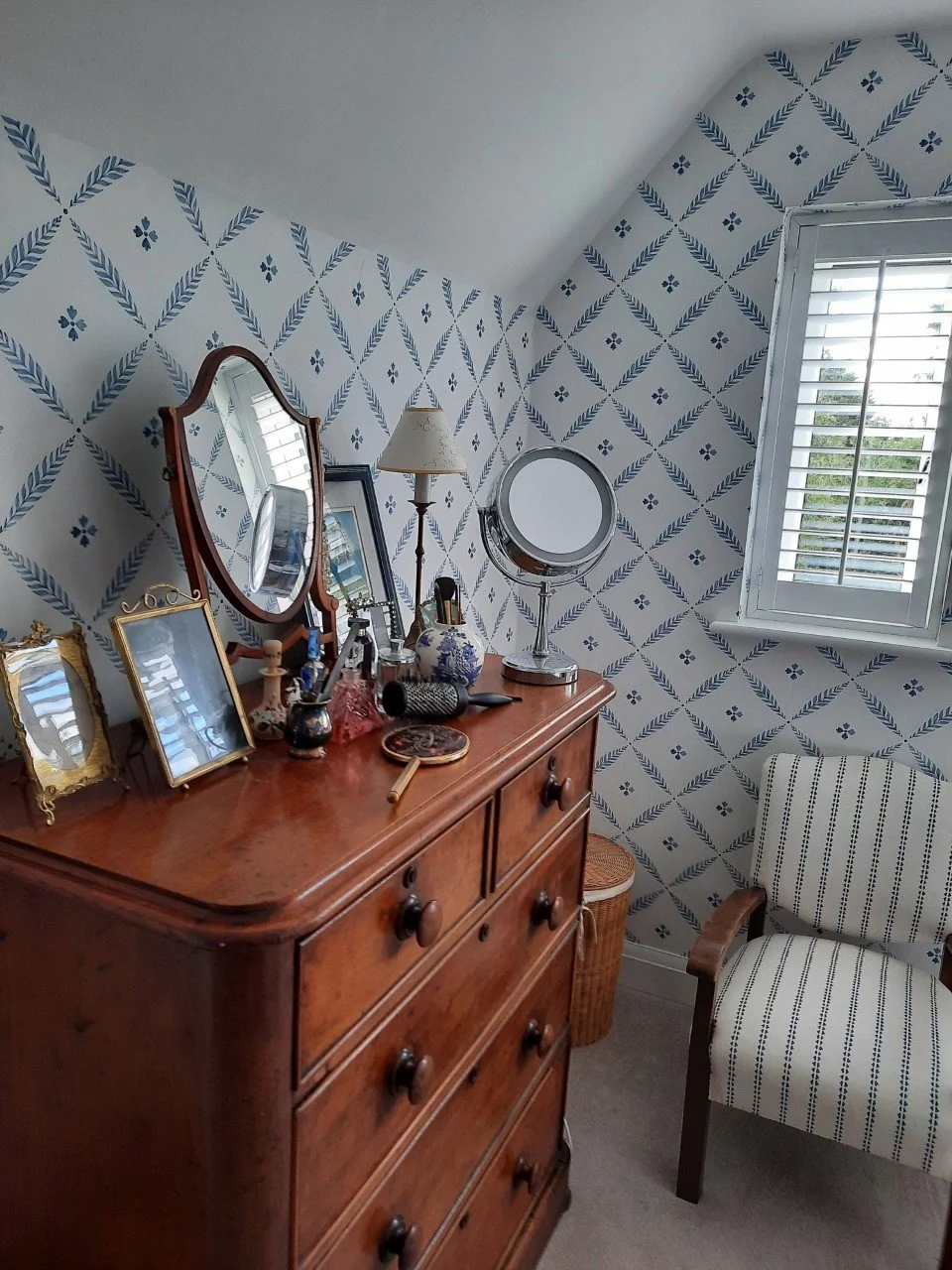 A bedroom corner with a wooden dresser topped with various small objects, mirrors, and a lamp. A striped upholstered armchair sits next to a window with white plantation shutters, and a wicker laundry basket is nearby. Blue and white patterned wallpa