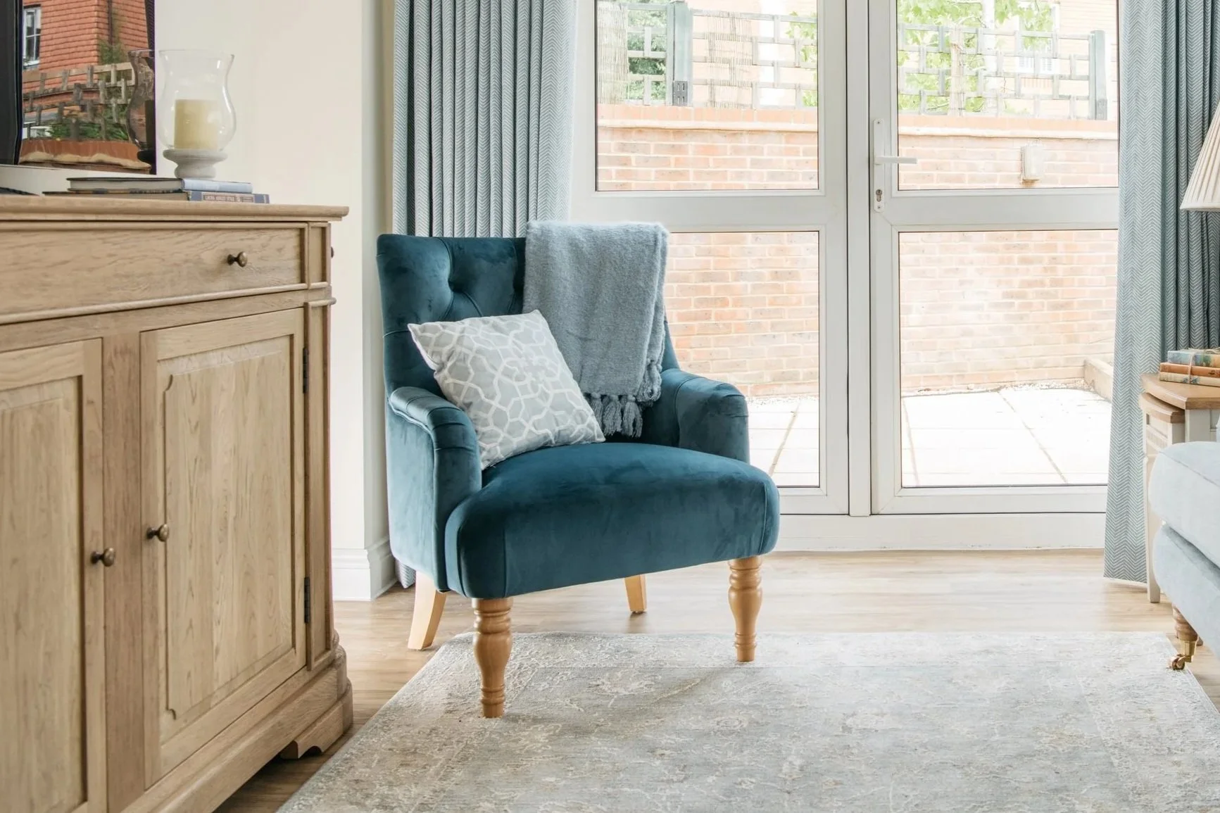 A living room with a blue armchair, decorated with a throw blanket and a pillow, next to a large window with curtains. A wooden cabinet with a candle and books is on the left side.
