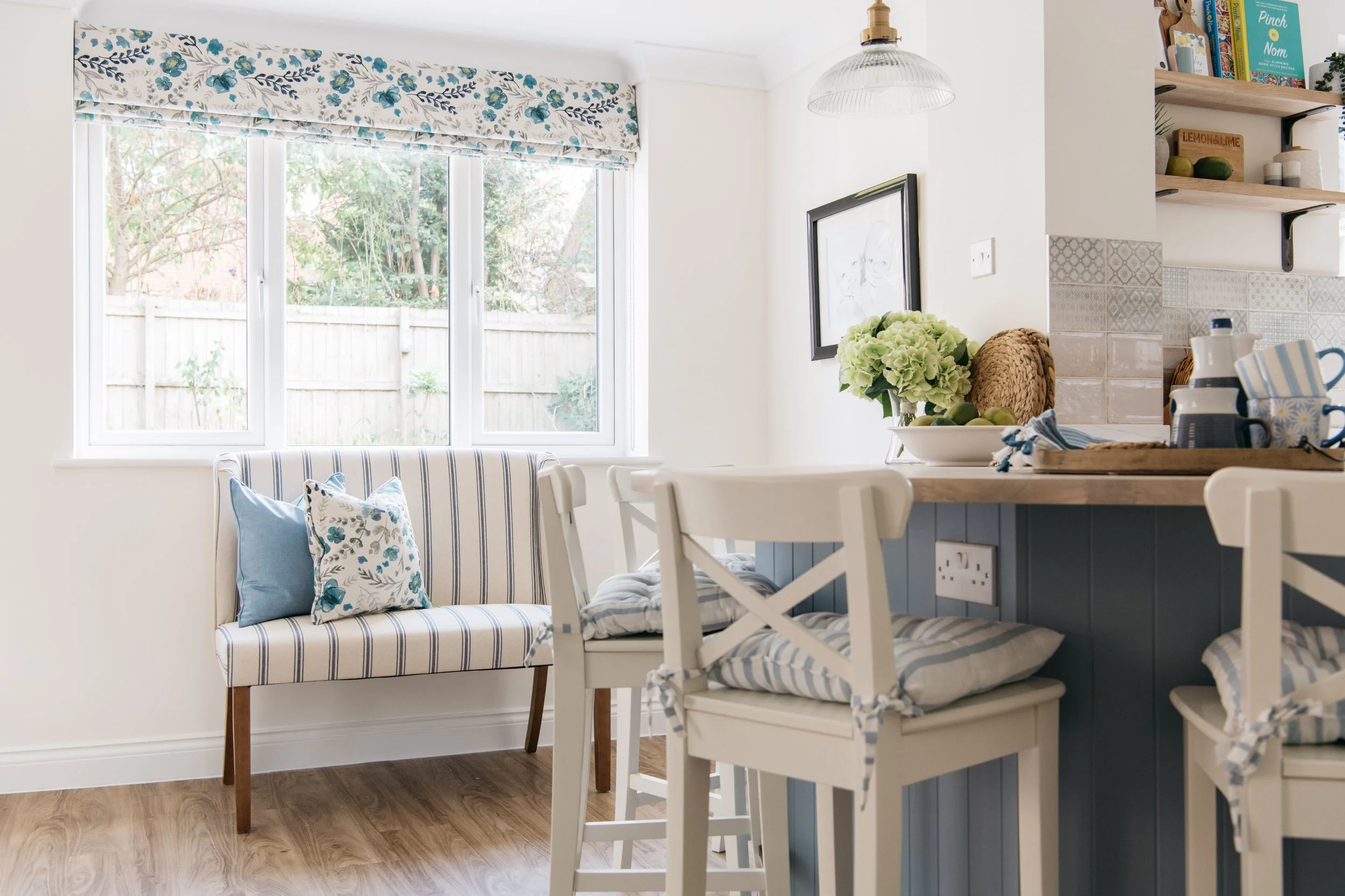 A bright kitchen and dining area with a window, a striped bench with blue and floral pillows, and a wooden bar with barstools, decorated in a coastal style with blue and white accents.