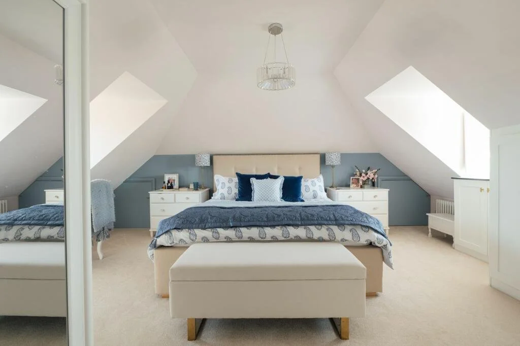 A large bedroom with a sloped ceiling and two skylights, featuring a large bed with blue and white bedding, white side tables with lamps and decorations, a beige bench at the foot of the bed, and a mirrored closet door.