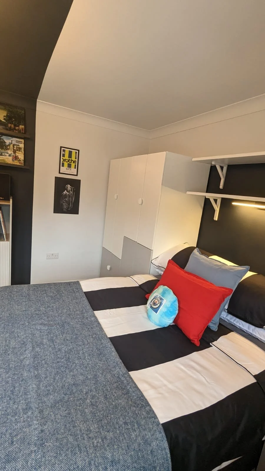 A teen bedroom with a bed that has a black and white striped bedspread, red and grey pillows There is a white wardrobe and wall shelves with a black wall behind the bed. The walls have picture frames and art, and a section of the ceiling is visible.