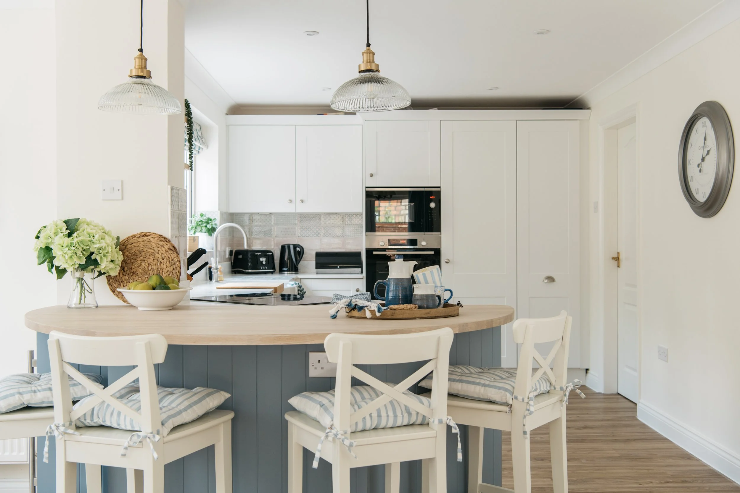Bright kitchen with white cabinets, a black oven, and a curved blue peninsula with custom oak breakfast bar seating. Decor includes a flower vase and bowls, with pendant lights hanging from the ceiling.