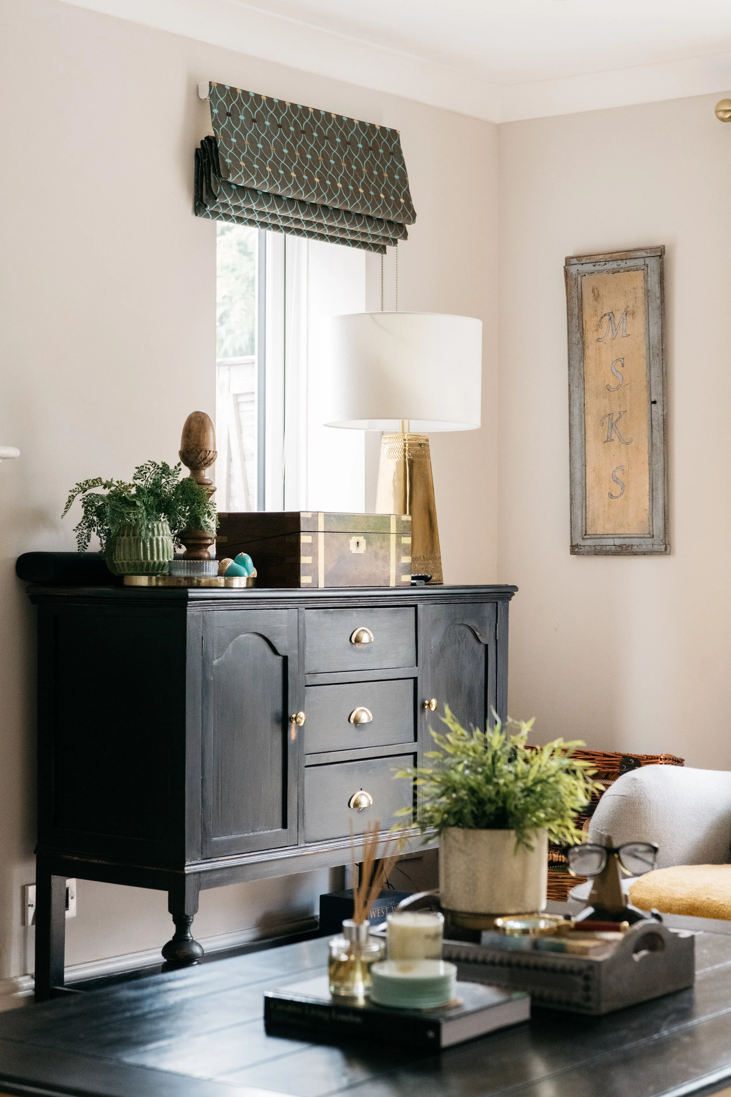 A living room corner with a black wooden sideboard, a white table lamp with a golden base, a framed art piece on the wall with 'MSK' letters, a window with a patterned shade, and a coffee table with decorative items and a plant.