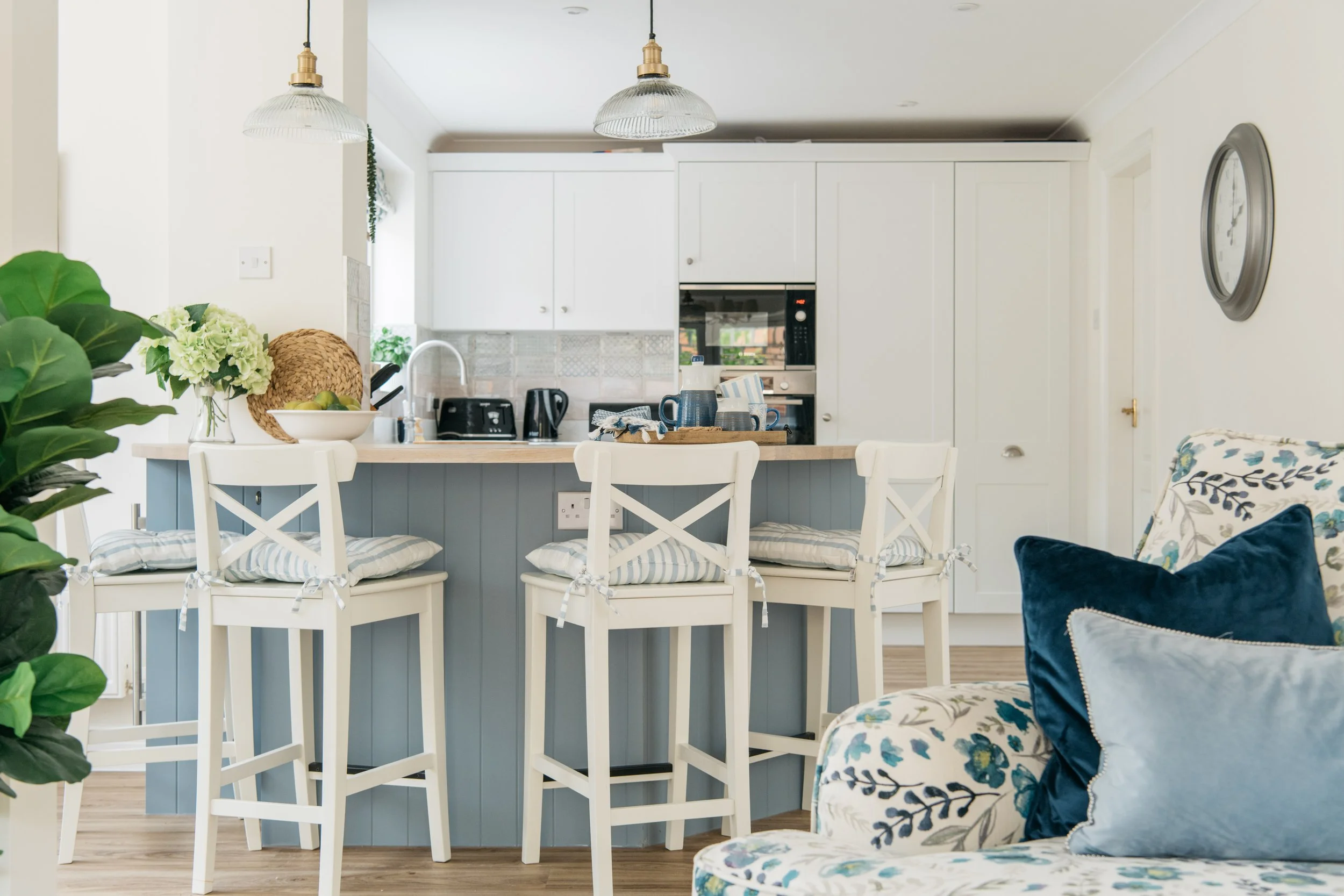 A bright, airy kitchen with white cabinetry and a light wood countertop. Three white bar stools with striped cushions are positioned at the kitchen island. The background features a sink, coffee maker, and microwave. To the left, a plant, a bowl with