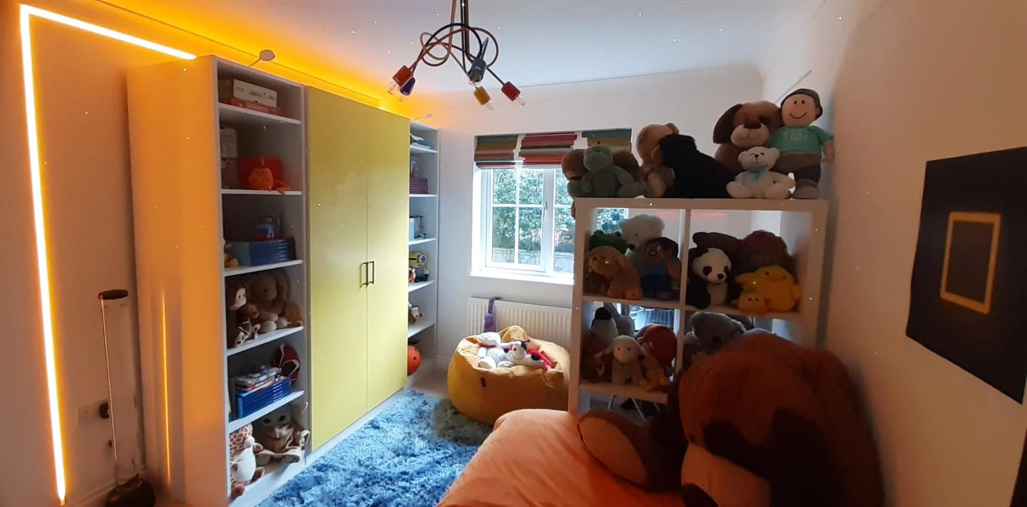 A cozy child's bedroom with a white bookshelf filled with stuffed animals and toys, a yellow wardrobe, a window with colorful striped blinds, an orange sofa with plush toys, and a fluffy blue rug There is a ceiling light fixture with colored bulbs an
