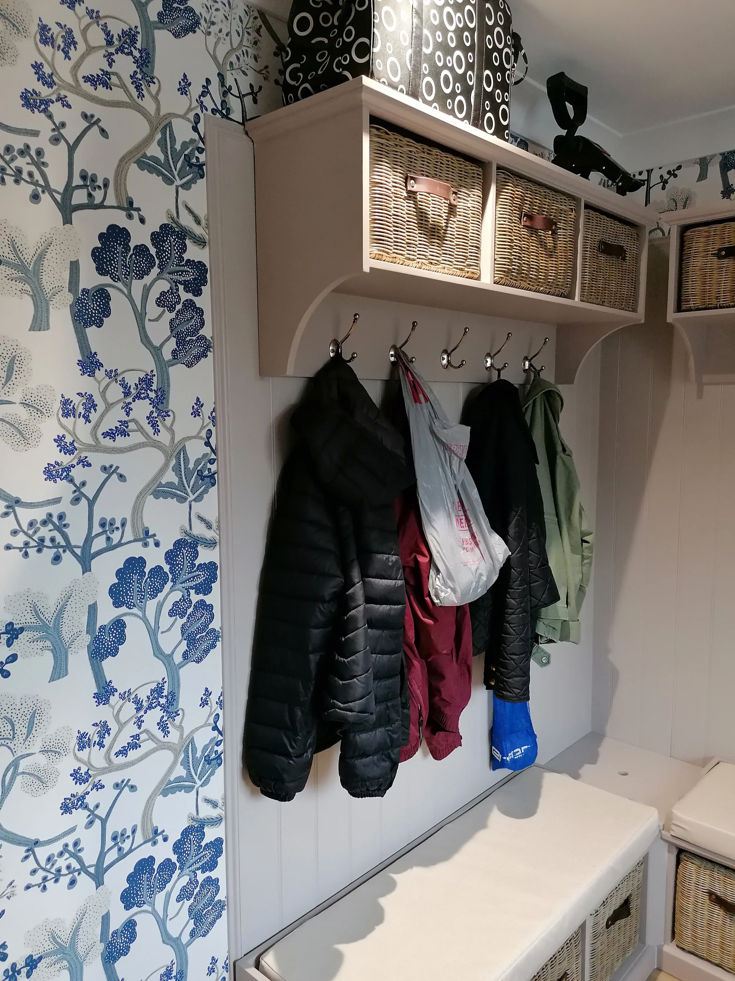 Boot room with coat hooks and wicker baskets on shelf, navy blue and white patterned wallpaper, and a white bench underneath.