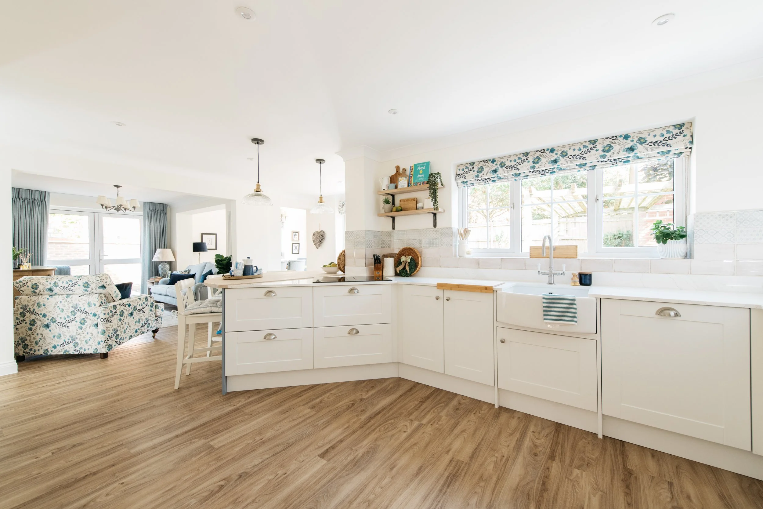 Bright, white kitchen with large window, wooden shelves, and white cabinets, opening into a living room with floral-patterned sofa and cozy seating area.