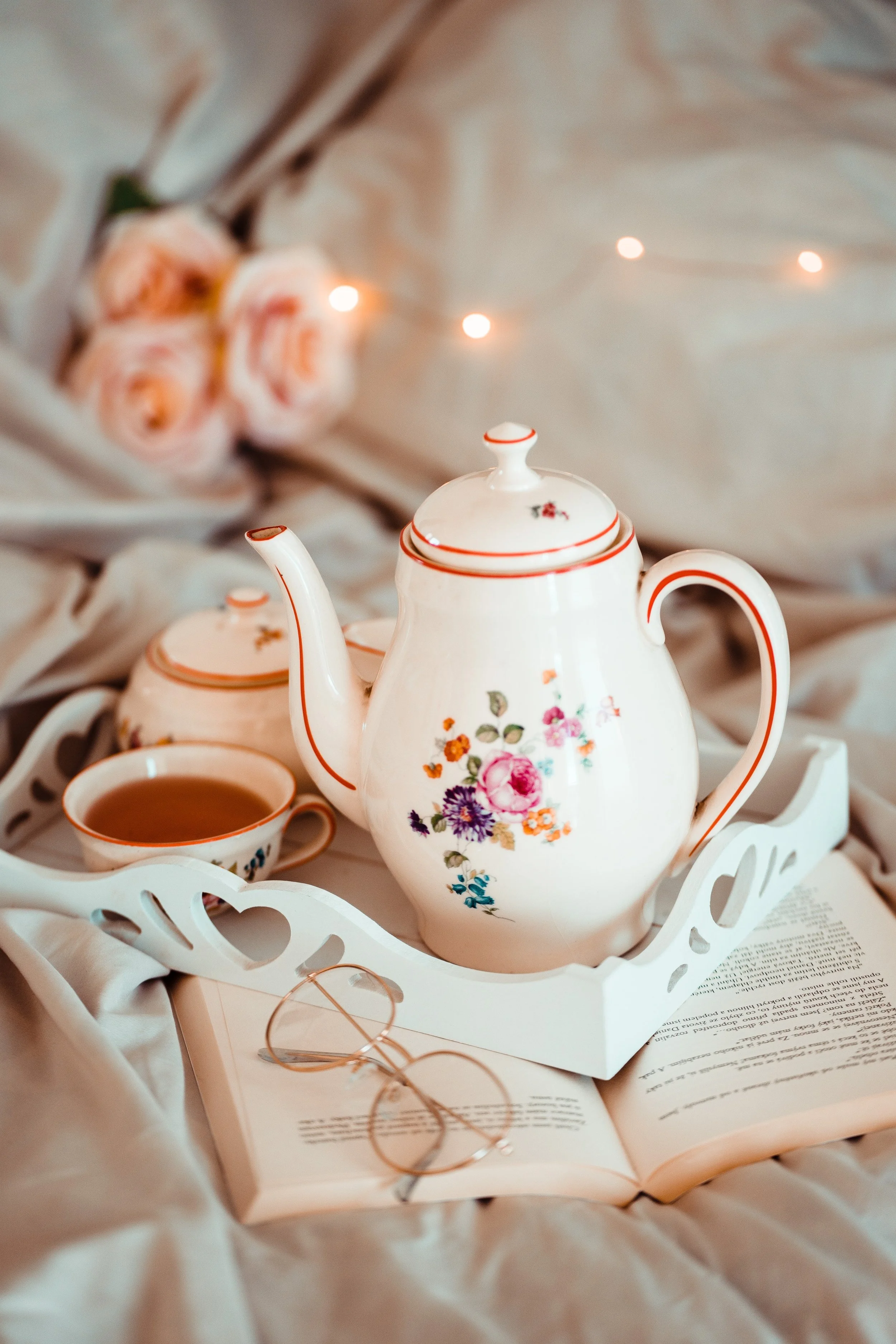 Pretty china teacup and teapot on a white tray with a book, flowers and fairy lights