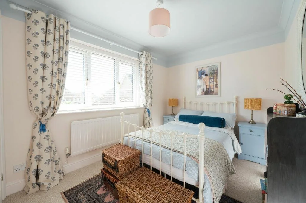 Bedroom with white iron bed, blue pillows, and beige bedding, two blue nightstands with yellow lamps, framed artwork above the bed, window with floral curtains, wicker storage baskets at the foot of the bed, and a black dresser with decorative items.