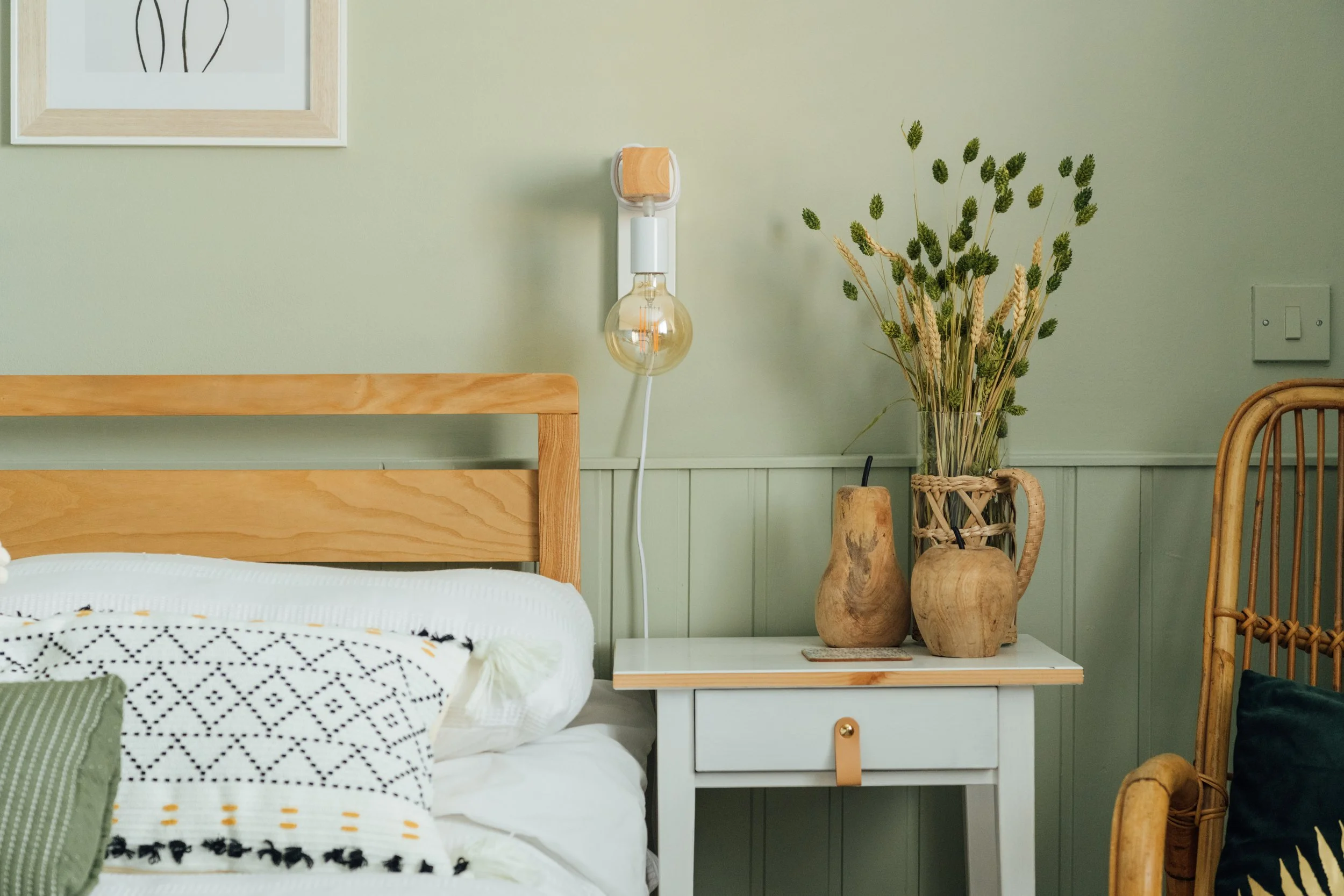 A bedroom corner with a wooden bed frame, a nightstand with decorative vases and dried flowers, and a wall-mounted lamp with a visible light bulb.