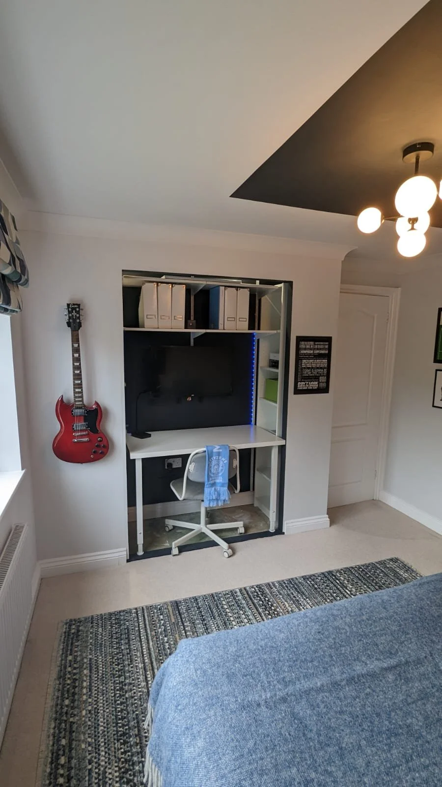 A bedroom corner with a small desk and a white office chair with a blue scarf draped over it. A red electric guitar is mounted on the wall next to the desk. There is a built-in shelf unit above the desk with storage boxes and a television. The room h