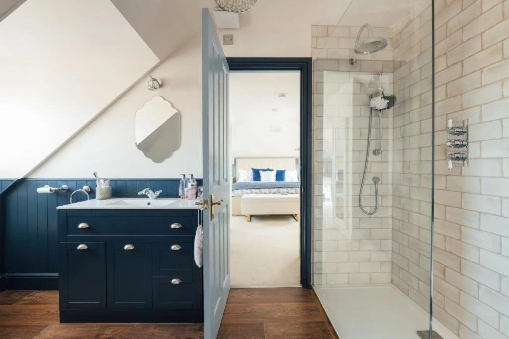 Modern bathroom with a walk-in shower and a vanity with a mirror, open to a bedroom with a bed and blue pillows.
