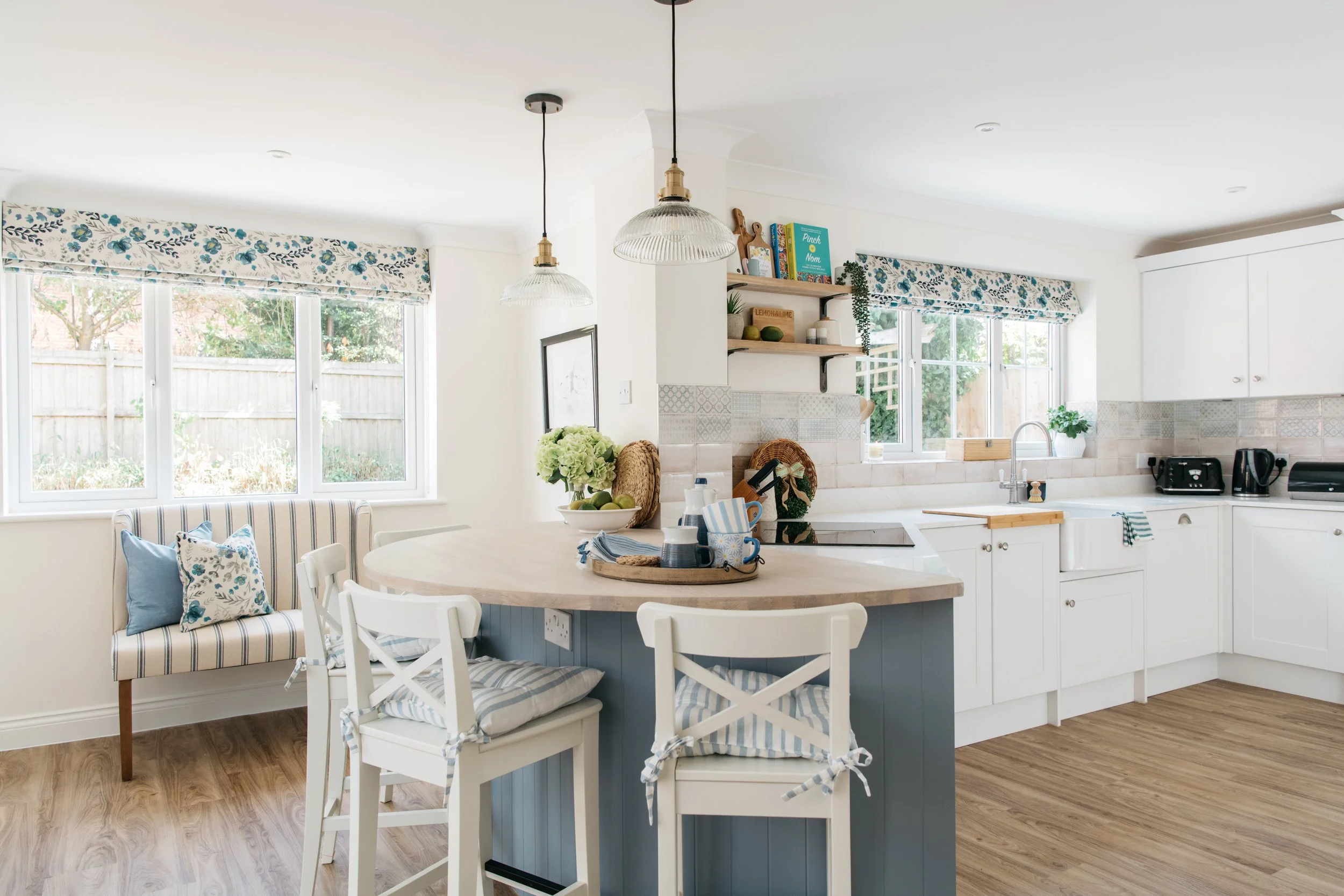 Bright kitchen with white cabinets, wooden countertops, and a blue island with seating. There are windows with floral valances, and shelves with decor and books. Decor includes a vase with flowers on the island and a few small appliances on the count