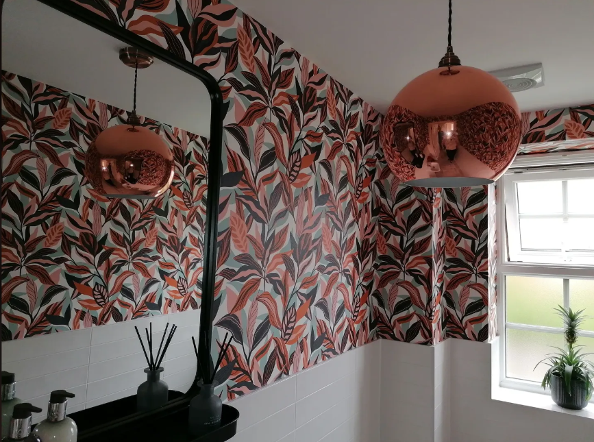 Interior of a room with a leafy patterned wallpaper in shades of pink, black, and beige, a large pink metallic pendant light, a window with a small green plant on the windowsill, and a mirror reflecting part of the room.