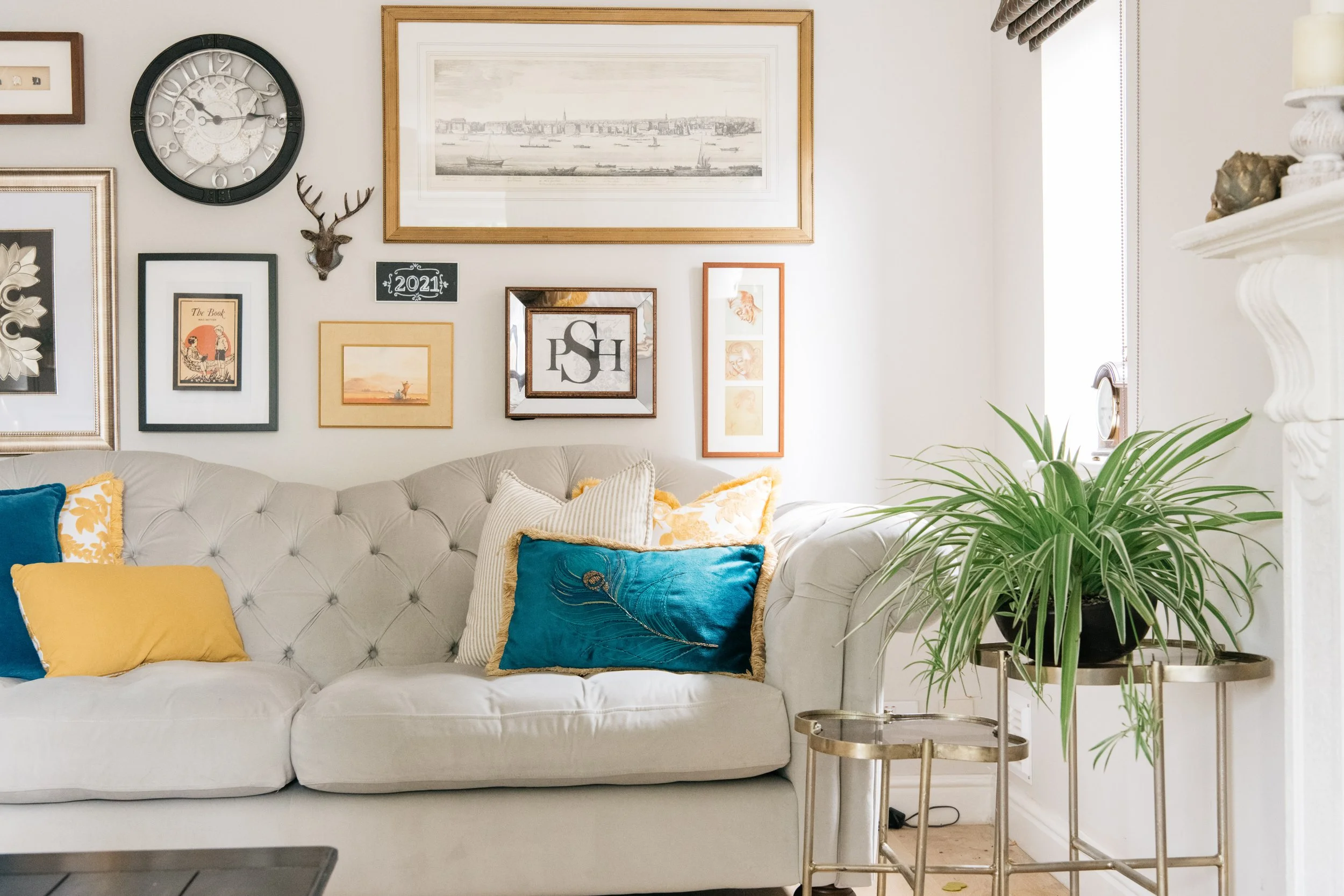 Living room with a beige tufted sofa decorated with colorful pillows and a gallery wall of framed artwork, a large green potted plant on gold stands, a window with blinds, and a white ornate fireplace mantel.