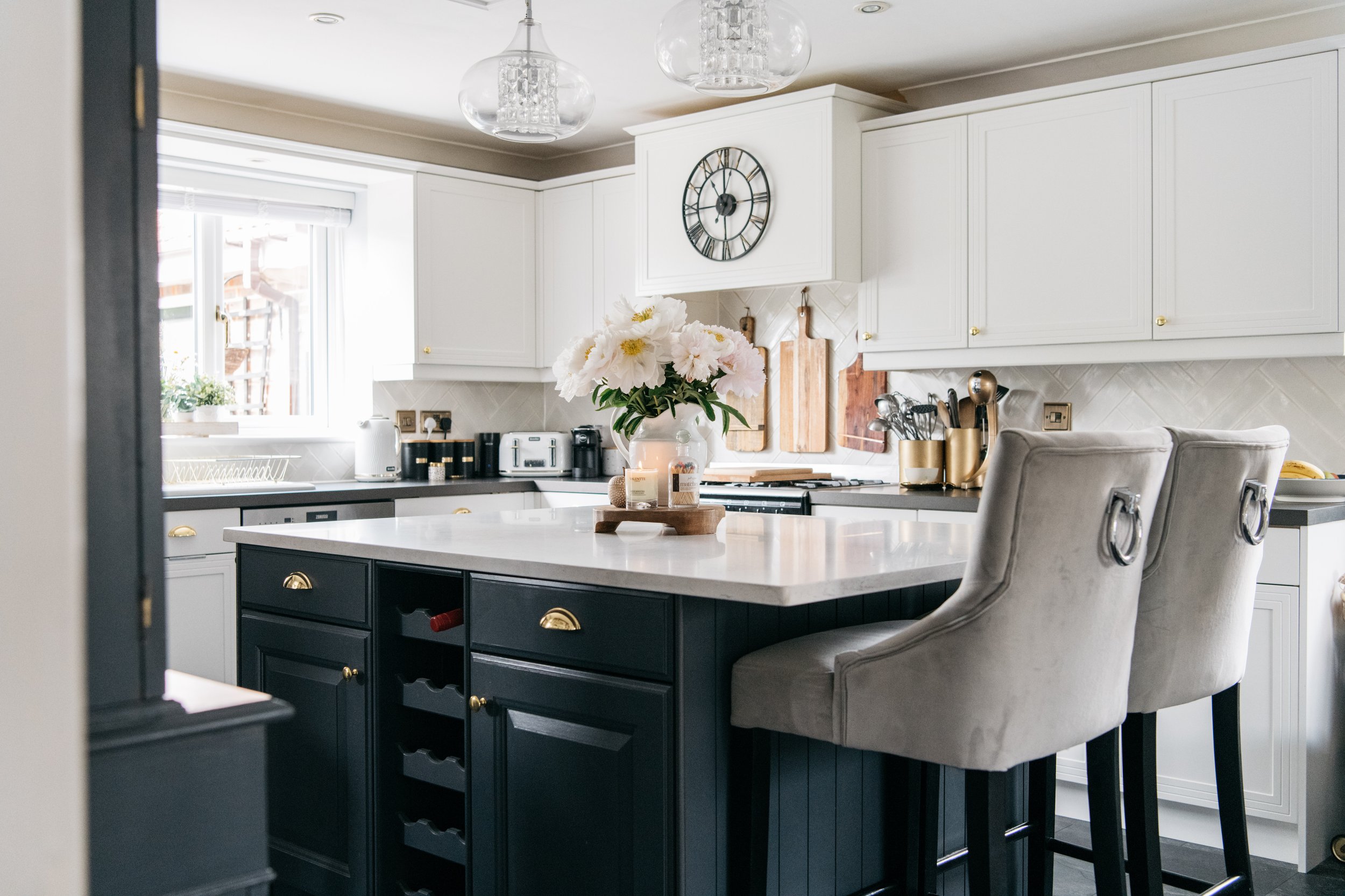 Modern kitchen with white cabinetry, a black island with gold handles, and a white countertop. There's a vase with flowers on the island and beige bar stools. A clock is mounted on the wall above the stove, and pendant lights hang from the ceiling.