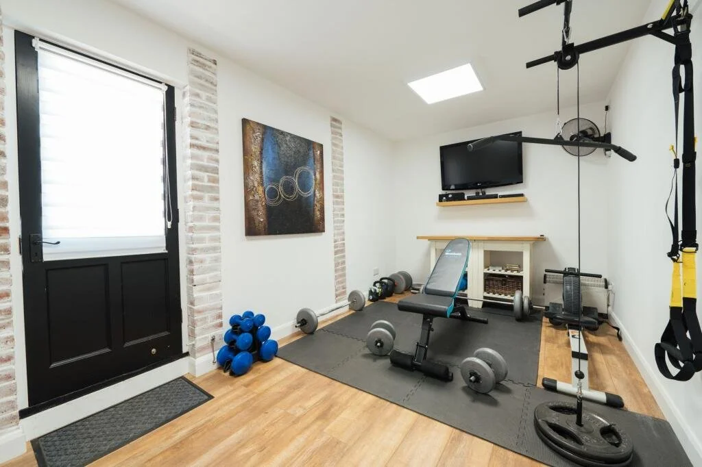Home gym with weights, a workout bench, resistance bands, a TV on the wall, and exercise equipment on a black rubber mat.