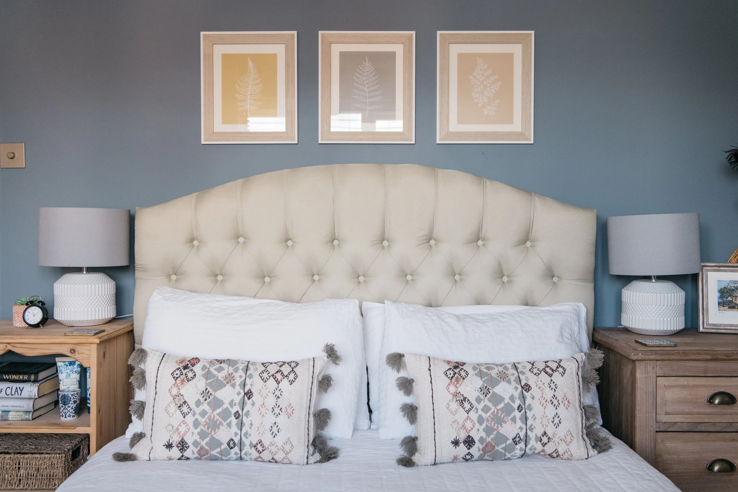 A neatly made bed with a tufted cream-colored headboard, two white pillows, and decorative pillows with geometric patterns and tassels, flanked by wooden nightstands with lamps, books, and framed photos, with three framed leaf prints on a blue wall a