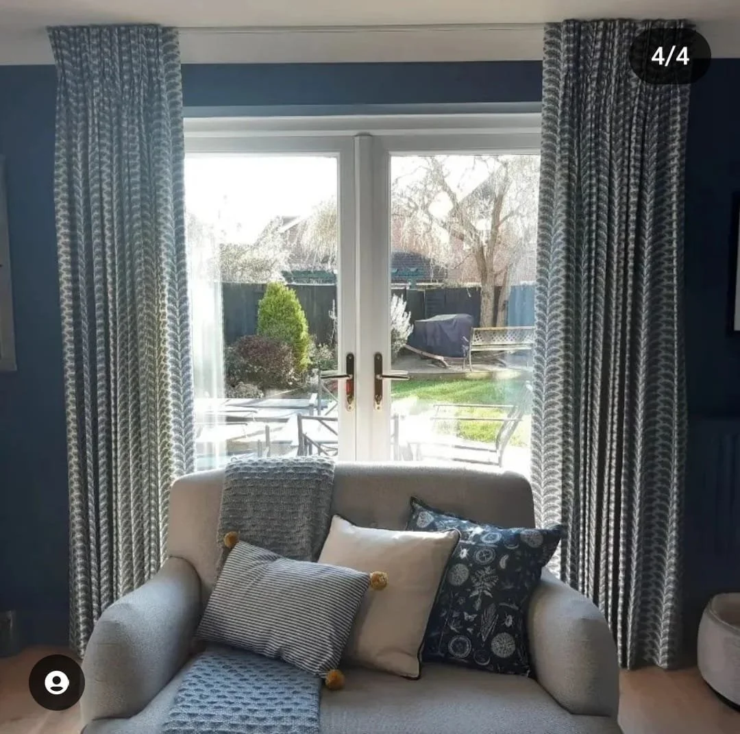 View of a living room with glass double doors leading outside, decorated with patterned curtains, and a comfortable sofa with multiple pillows in front of the doors.