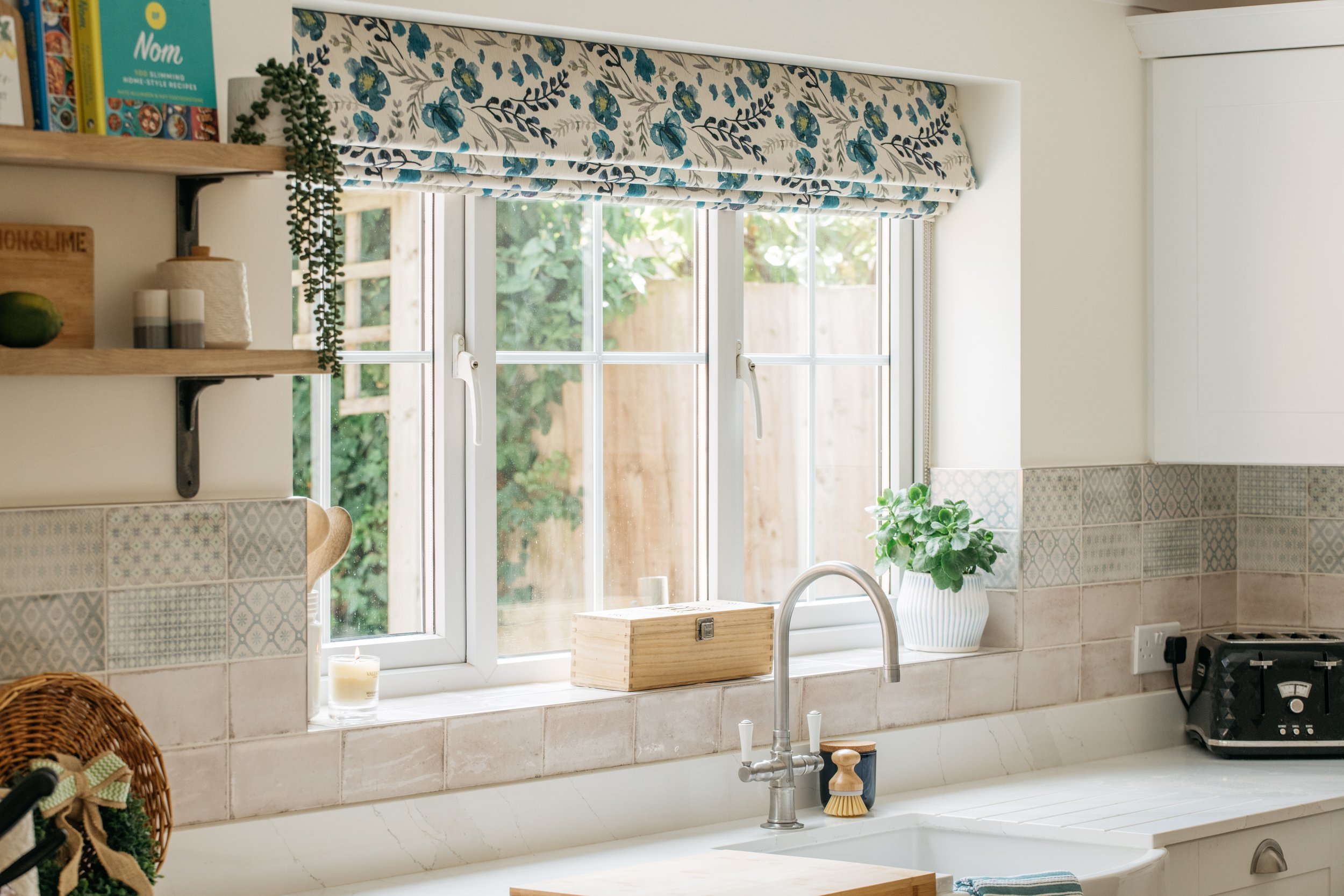 Kitchen window with a bespoke Roman blind, potted plant, wooden box, candle, toaster, and sink with faucet.