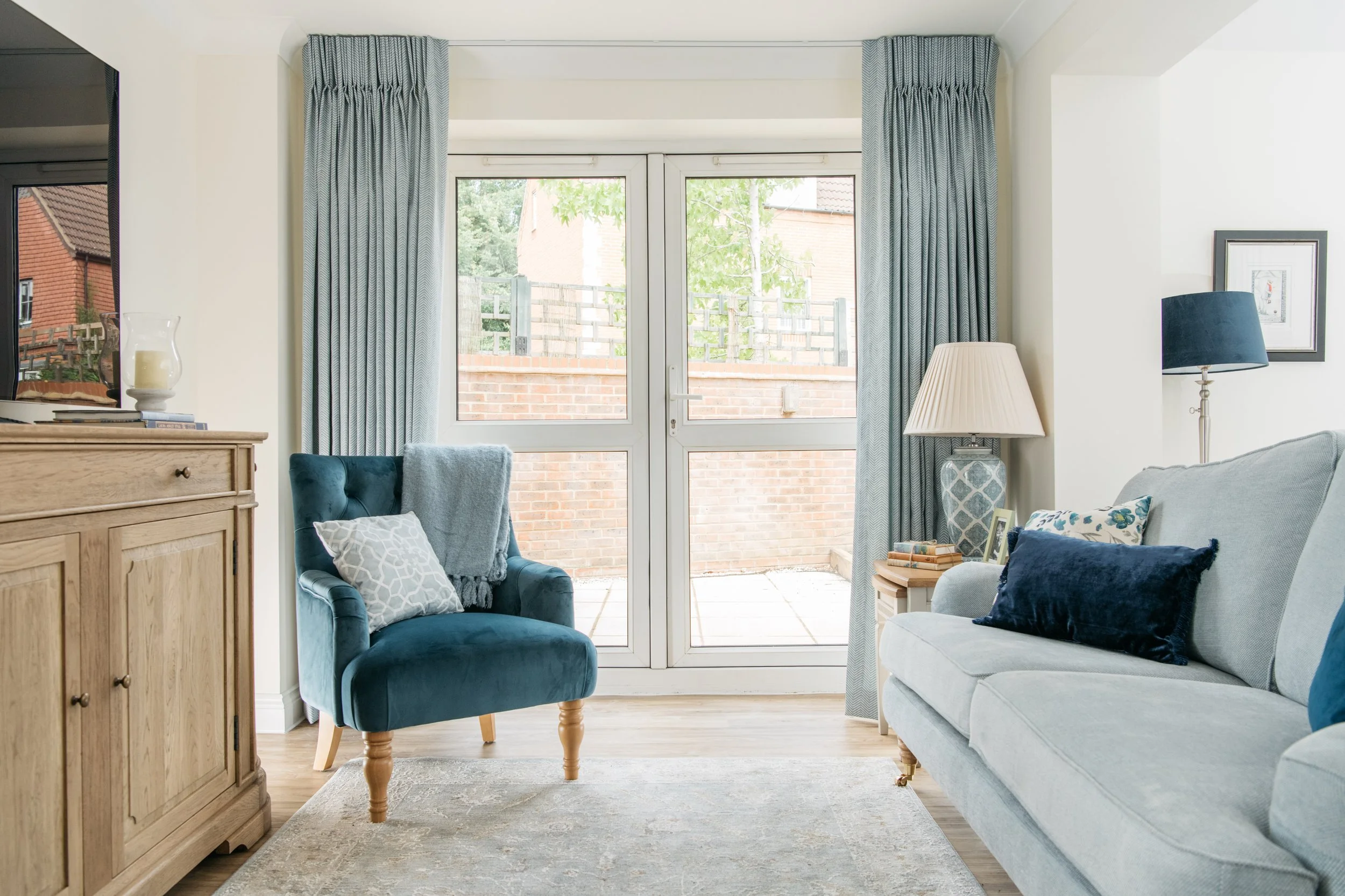 Family area with light grey sofa, blue armchair, wooden side table with a lamp and books, large glass sliding doors with light blue curtains, and outdoor patio area.