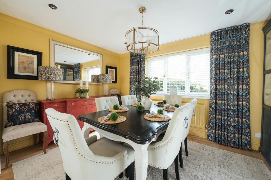 Yellow dining room with six white upholstered chairs around a dark wooden table set for a meal, a red sideboard, large window with patterned curtains, and decorative items including a mirror, lamps, candles, and a plant.