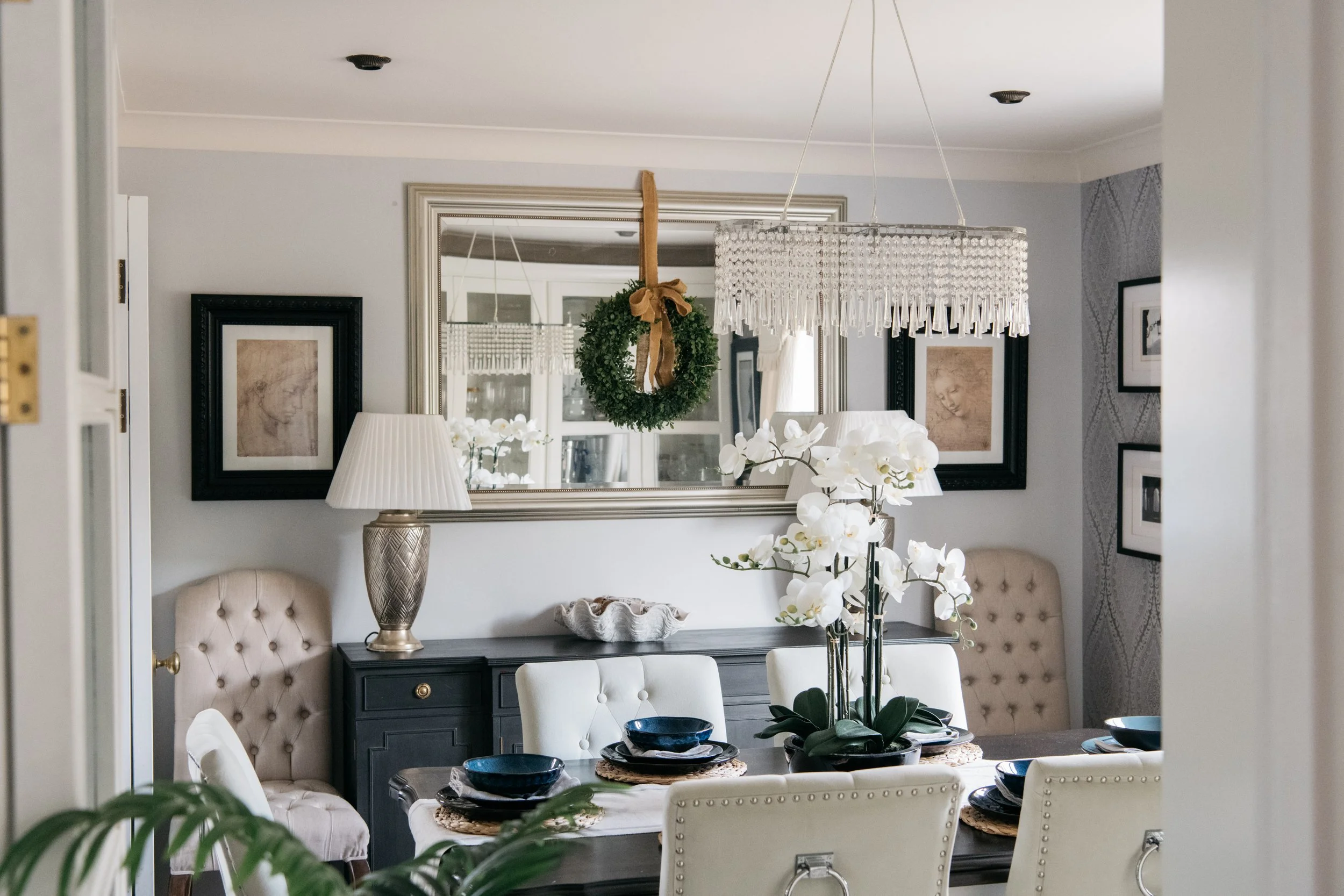 A dining room with a dark wooden table set with blue bowls and black plates, surrounded by white upholstered chairs with silver nailhead trim. A large vase with white orchids is in the center of the table. A black sideboard with a beige decorative bo