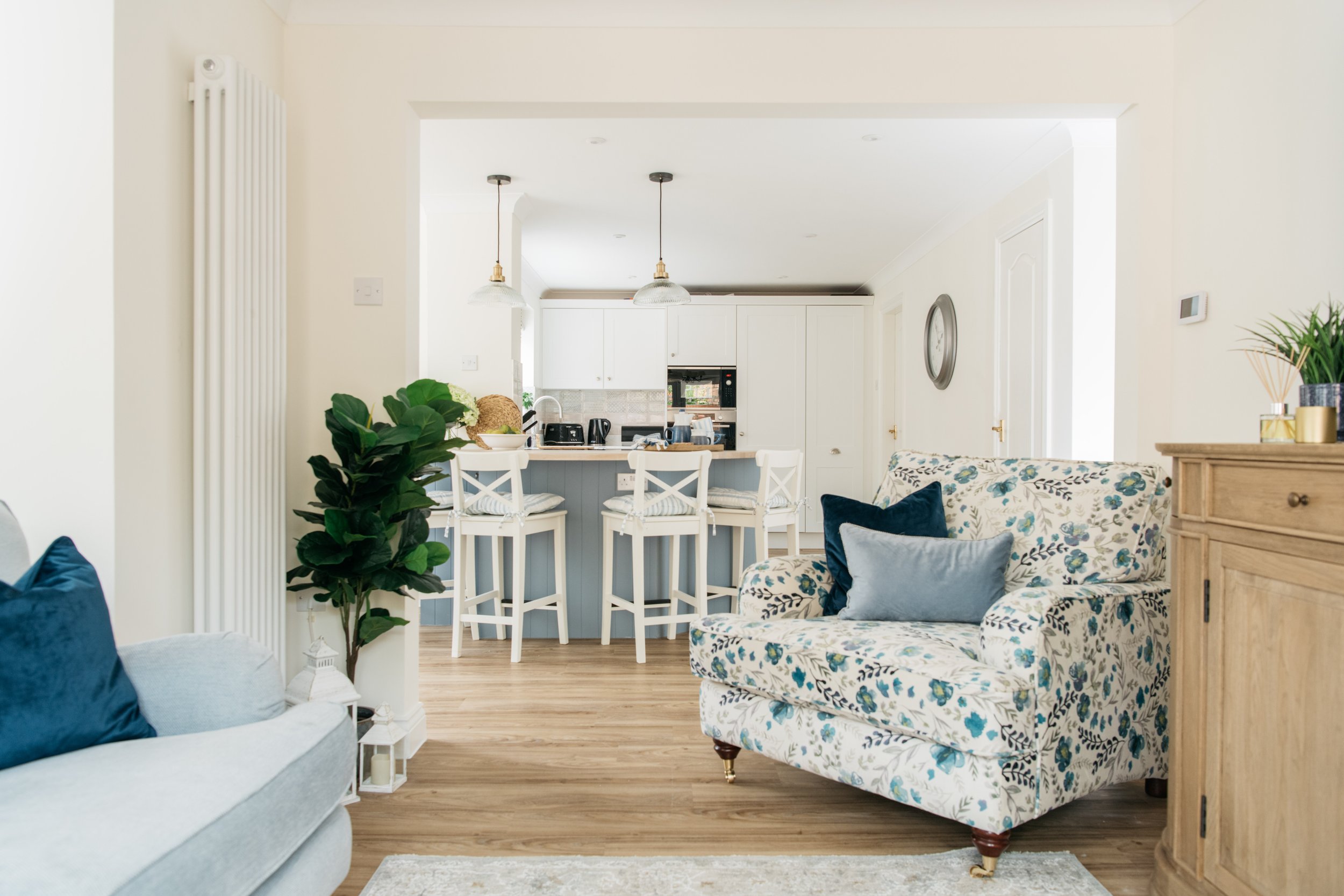 Living room and kitchen area in a home, featuring a floral upholstered sofa with decorative pillows, a light-colored armchair with a navy pillow, a wooden sideboard with small plants, a large green potted plant, and a breakfast bar with four white ch