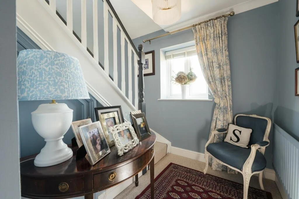 A cozy loft entryway with a staircase, a window with floral curtains, a small bench with a pillow, and a console table displaying family photos and a table lamp.