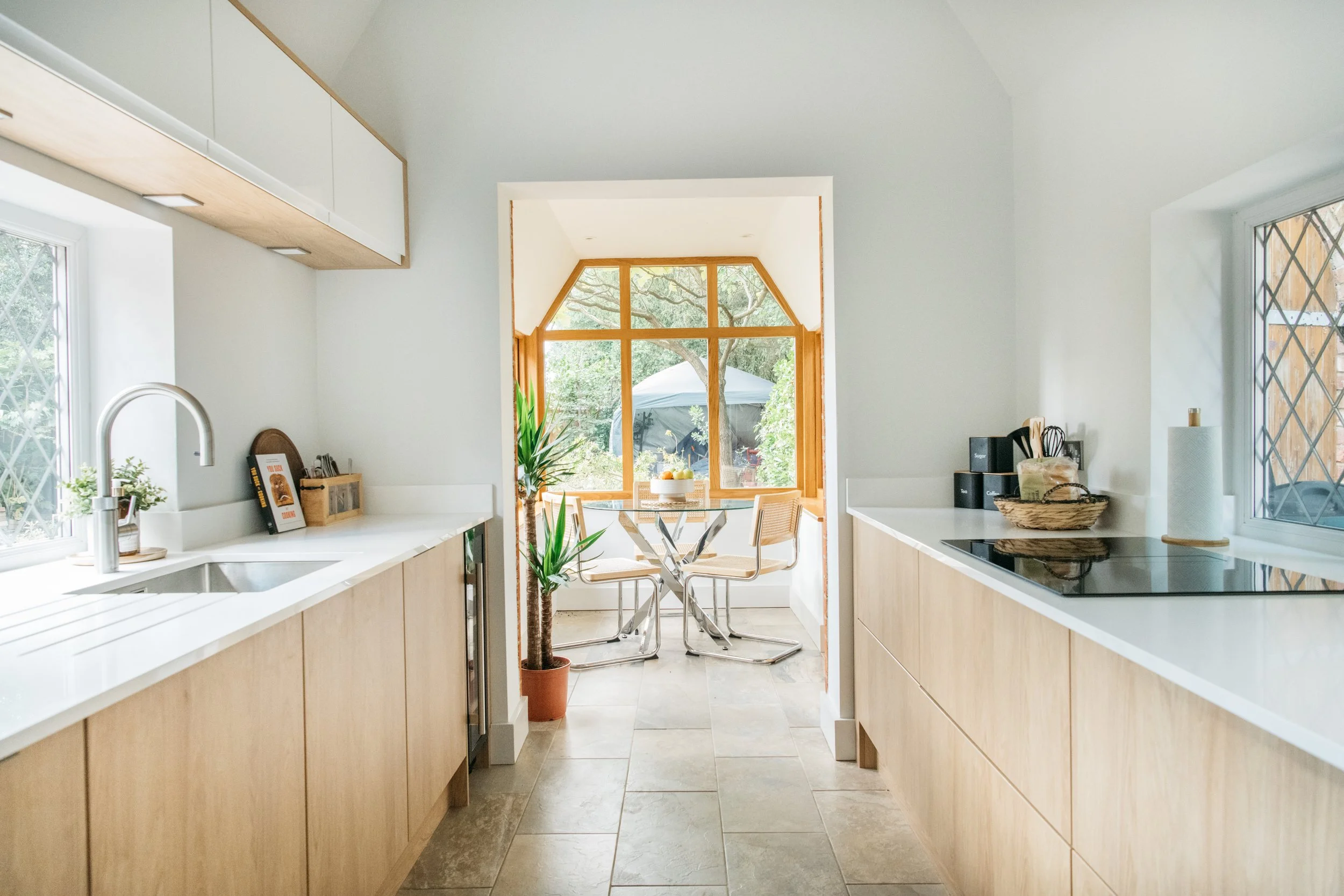 Bright kitchen with light wood cabinets, white countertops, and a view of a garden dining area through a large window with wooden trim.