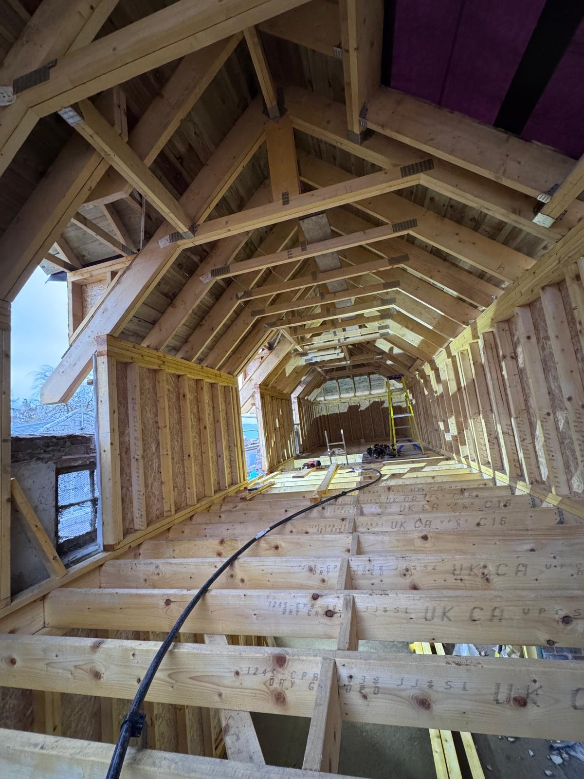 Interior view of a wooden attic space under construction, showing the exposed framework, rafters, and plywood flooring, with construction tools and equipment.