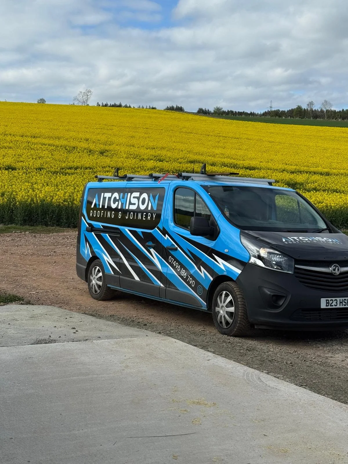 A blue and black advertising van parked on a dirt road in front of a yellow flowering crop field, with clouds and trees in the background.