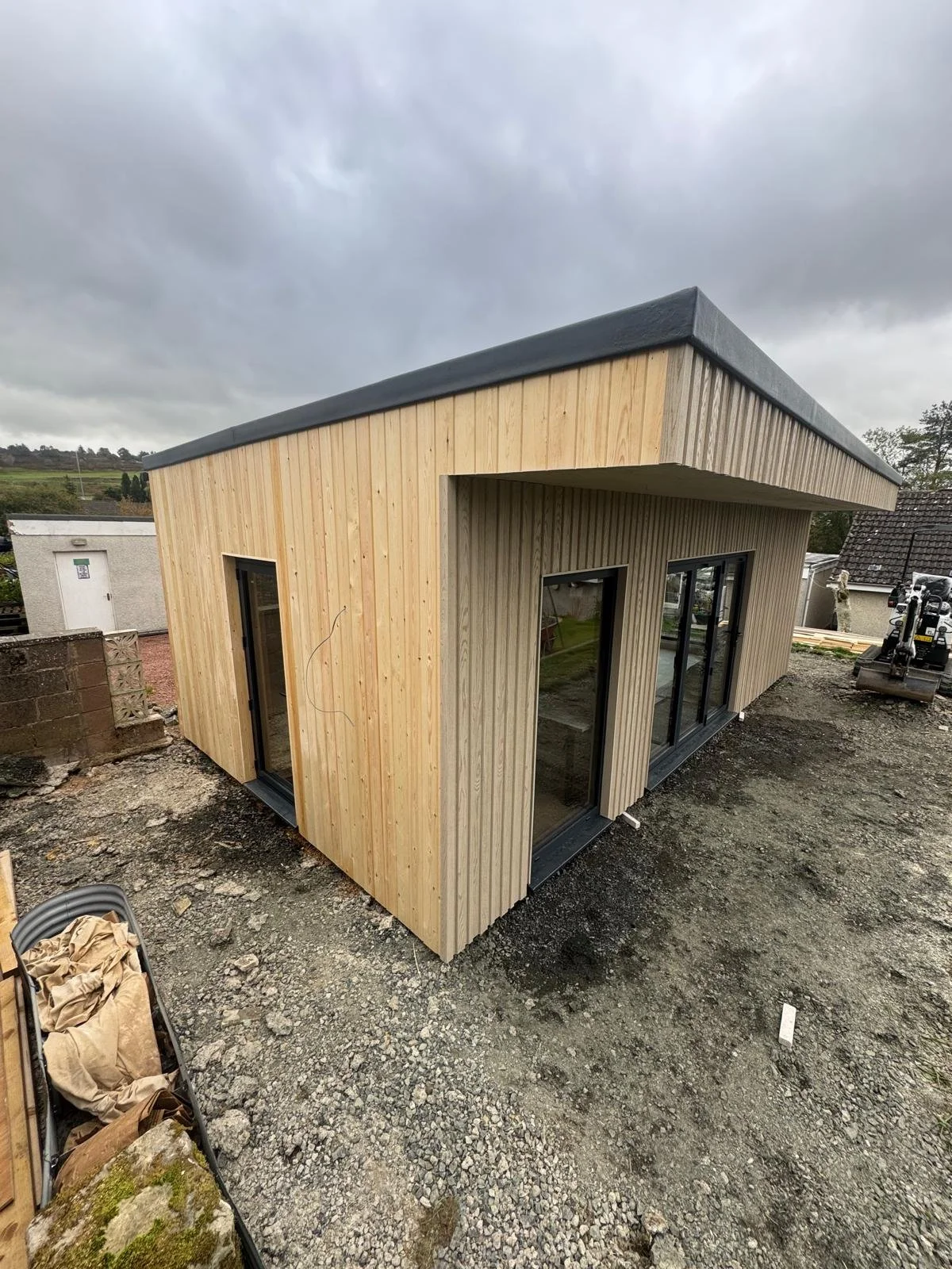 A small modern building under construction with light wood vertical siding, large glass sliding doors, and a slightly sloped roof. The construction site has loose gravel and dirt ground, and construction equipment is visible on the right. Garden room