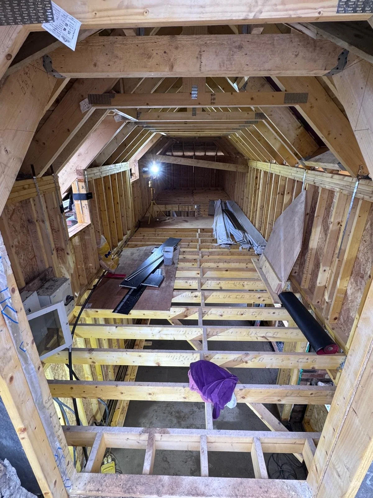 The interior of a house under construction with exposed wooden framing, some construction tools, and materials. The roof structure is visible, and there is a window opening on the left side.