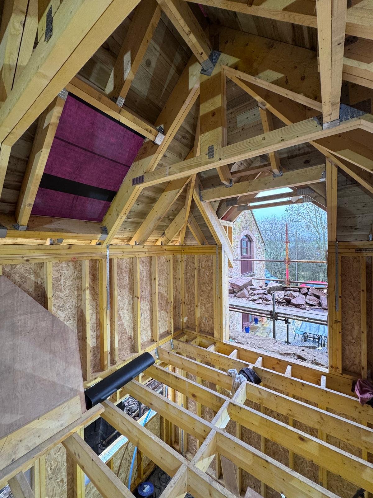 Inside view of a house under construction with wooden framing, insulation, and large window opening showing the outside yard and neighboring house. extension in scottish borders