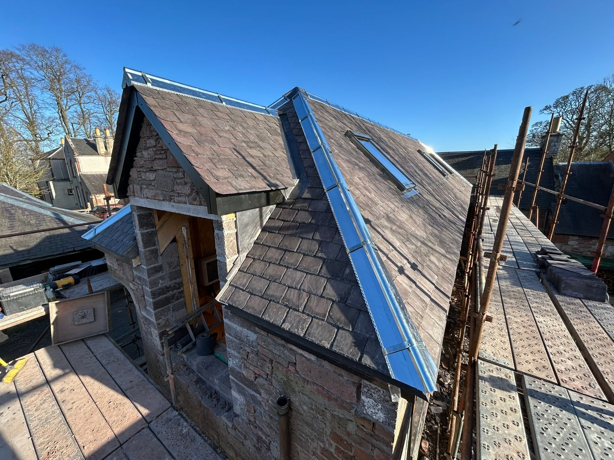 Construction site on a rooftop with scaffolding, shingles, and skylights installed on a sloped roof under a clear blue sky. new roof in scottish borders