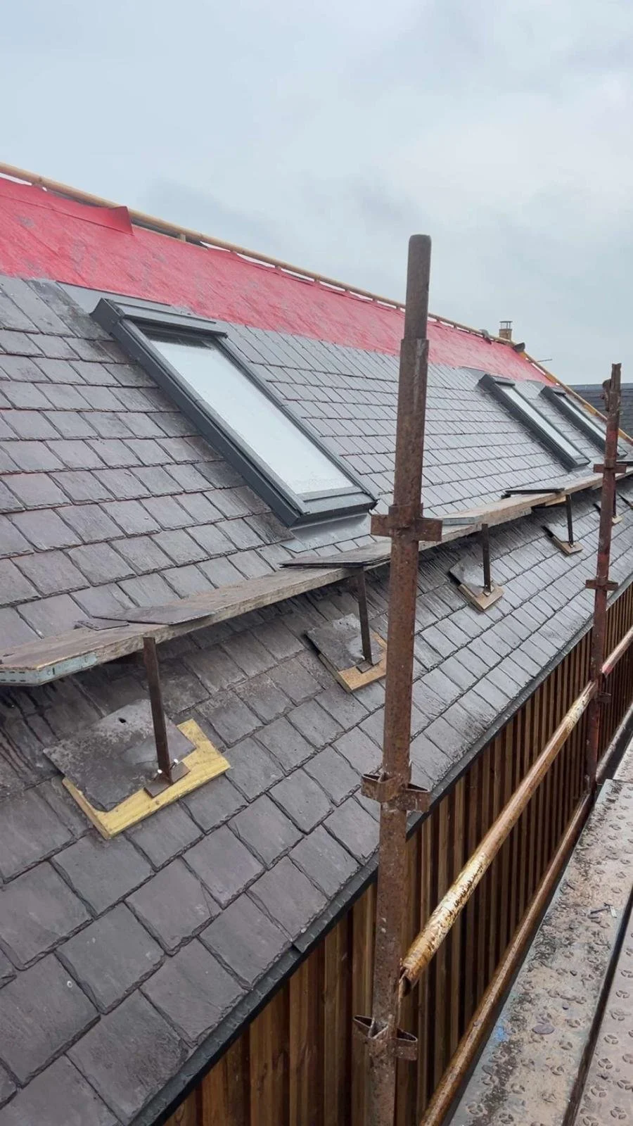 Construction scaffolding on a roof with two skylights, some roofing tiles, and a section of red underlayment.