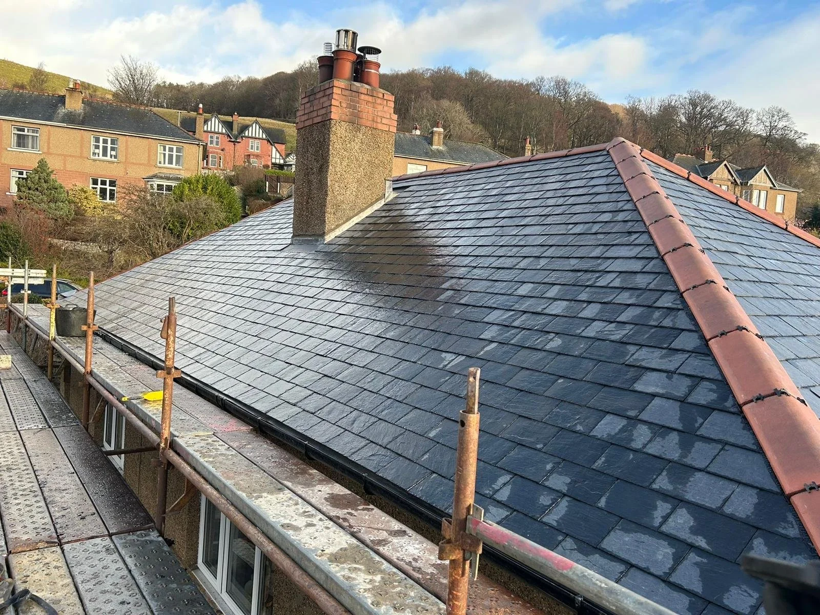 A house rooftop with slate shingles and a brick chimney, with neighboring houses and hills in the background under a partly cloudy sky.