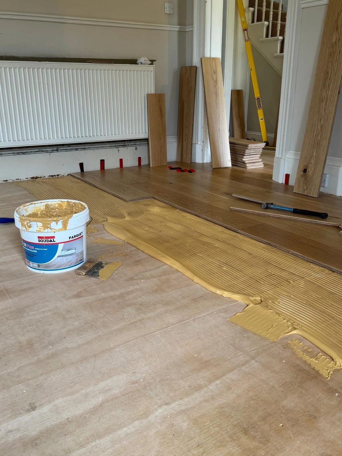 Room with hardwood flooring being installed, with a bucket of wood floor adhesive, wooden planks, a level, and a saw on the floor. The wall has a white radiator, and there are several wooden boards leaning against the wall. A yellow ladder is visible