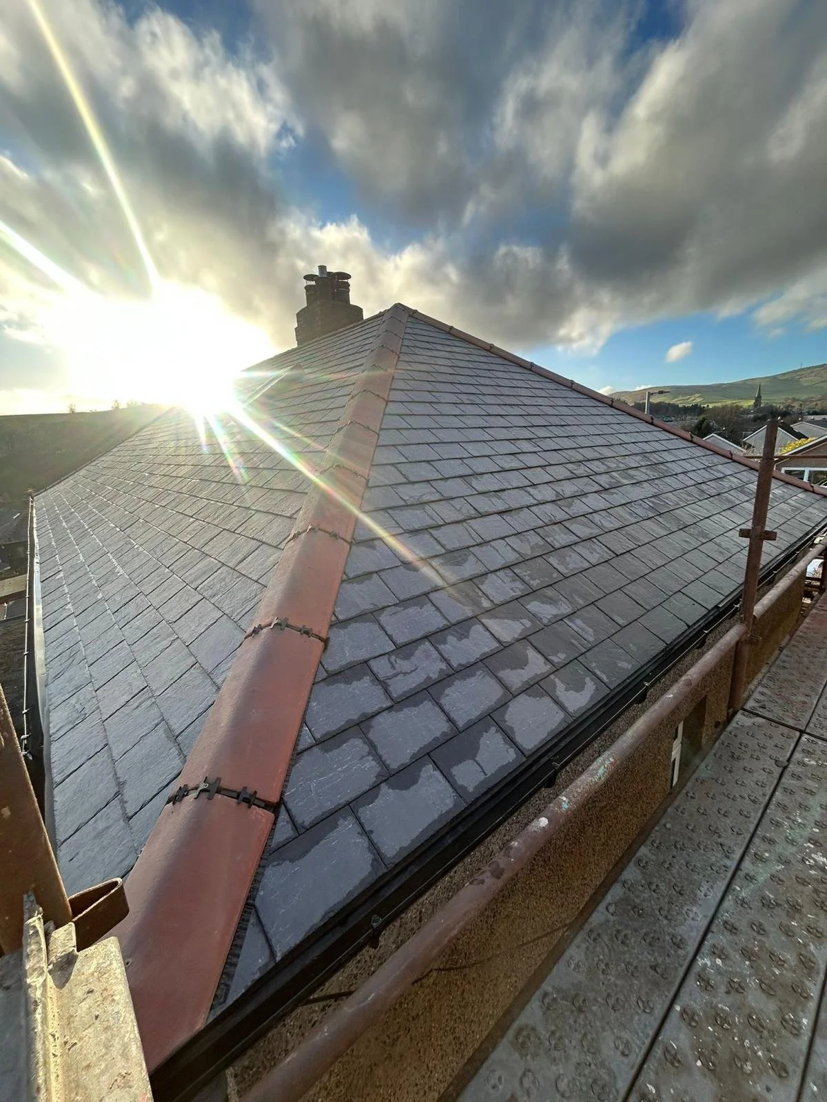 The image shows a roof with new gray tiles, a red ridge cap, and a chimney in the background. The sun is shining from the left, creating lens flare and illuminating the sky with clouds and blue patches. Safety scaffolding surrounds the roof.