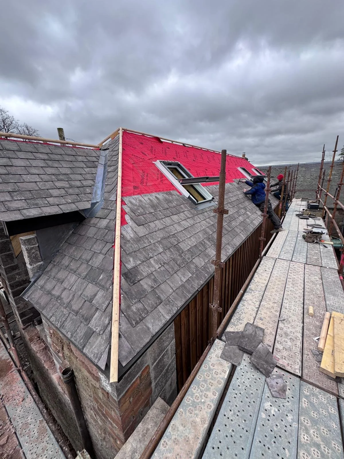 Construction workers installing roof windows on a sloped roof with gray shingles, scaffolding surrounding the building, and a cloudy sky overhead.