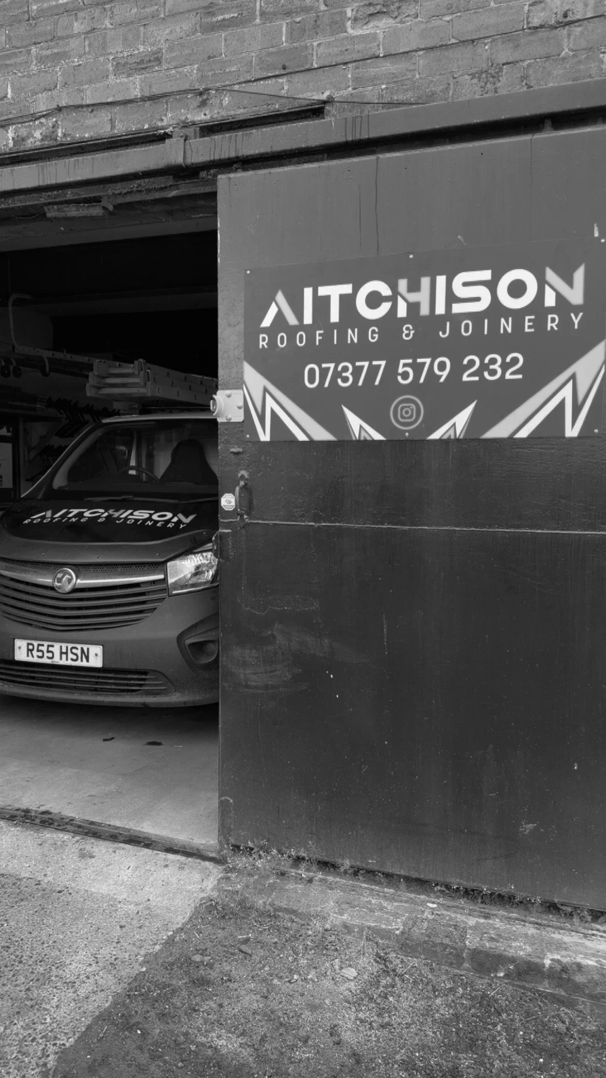 A vehicle parked inside a garage next to a sign that reads "Aitchison Roofing & Joinery" with a phone number and social media icon, viewed through a partially open garage door.