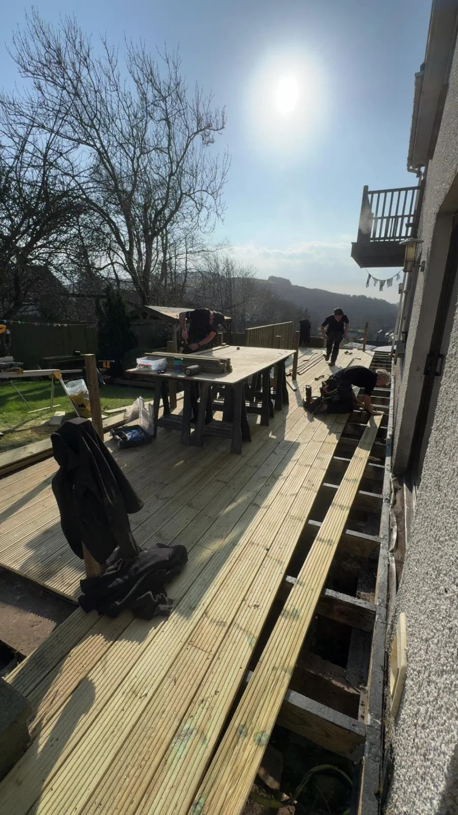 People working on deck construction, installing wooden flooring, with tools and materials on a scaffolded outdoor deck during daytime under a clear sky.