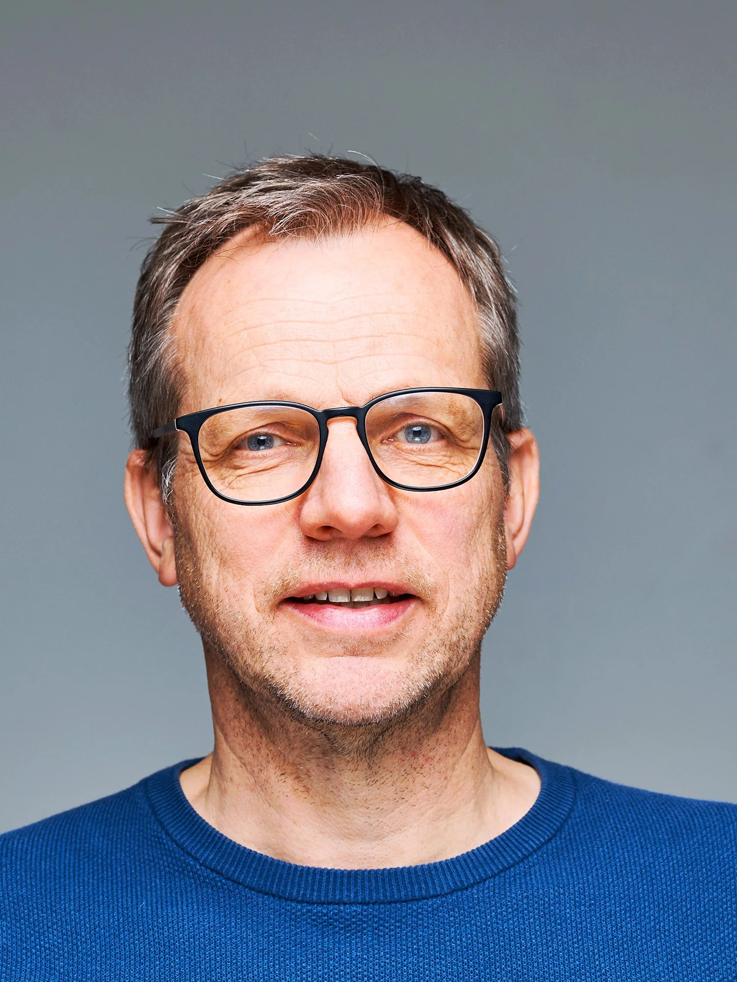 Nahaufnahme eines Mannes mit bräunlich-grauem Haar, blauen Augen und Brille, lächelnd. Trägt ein dunkelblaues Hemd vor einem grauen Hintergrund.