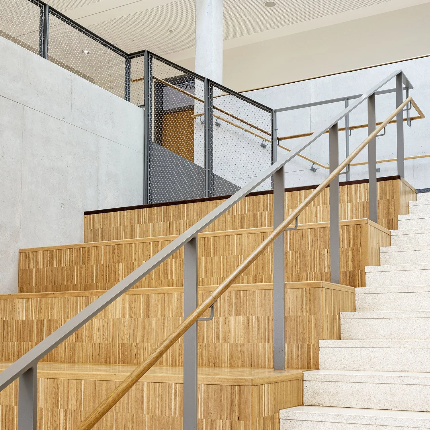 Innenaufnahme einer modernen Treppe mit Holz- und Betonelementen, geländer aus Metall mit Holzbalken, im Hintergrund eine weiße Wand und eine Metallabsperrung.