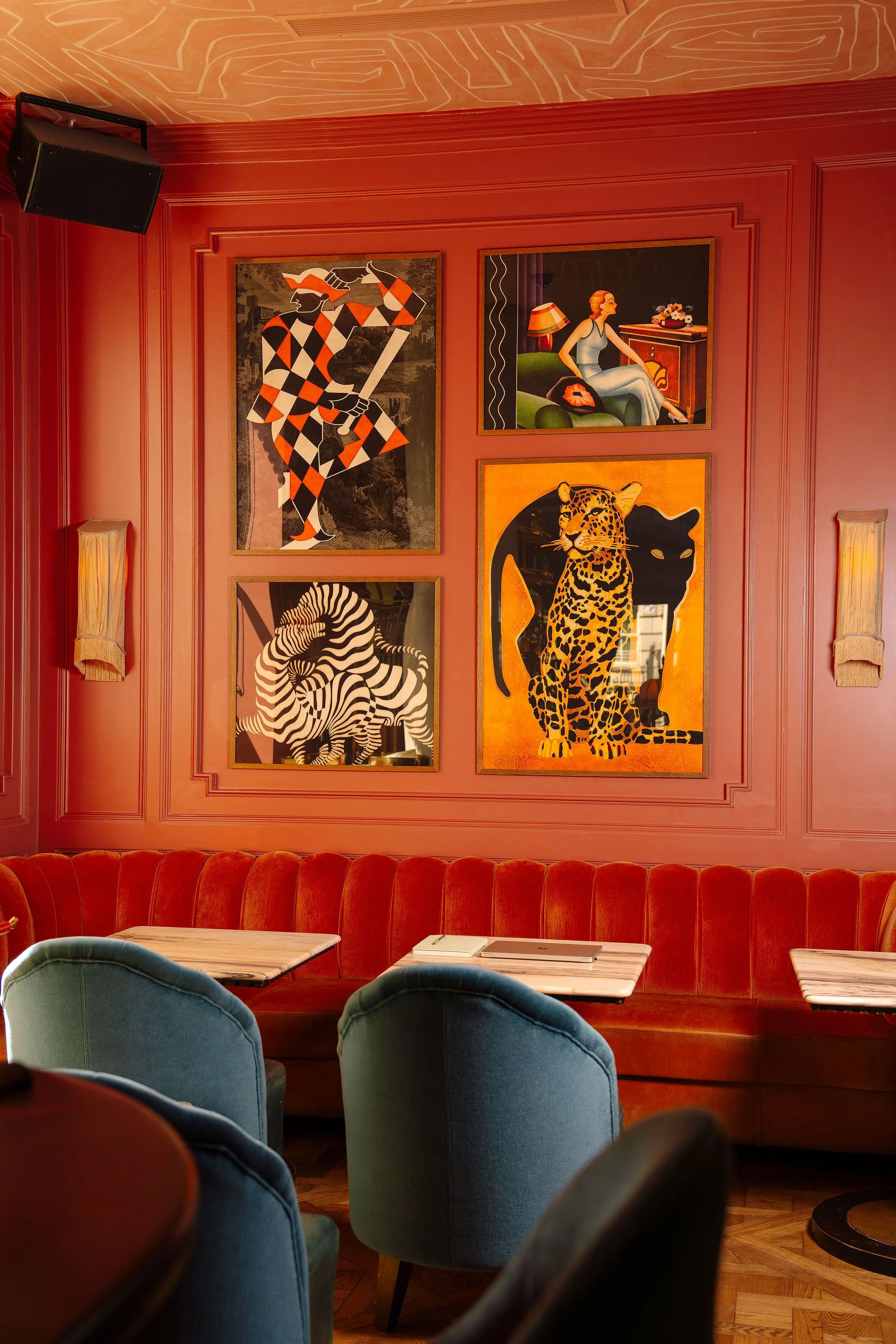 A cozy dining area with a red upholstered banquette, blue chairs, and tables with books and laptops. The wall behind features modern art paintings of animals and a woman, set against pinkish-red wall paneling.