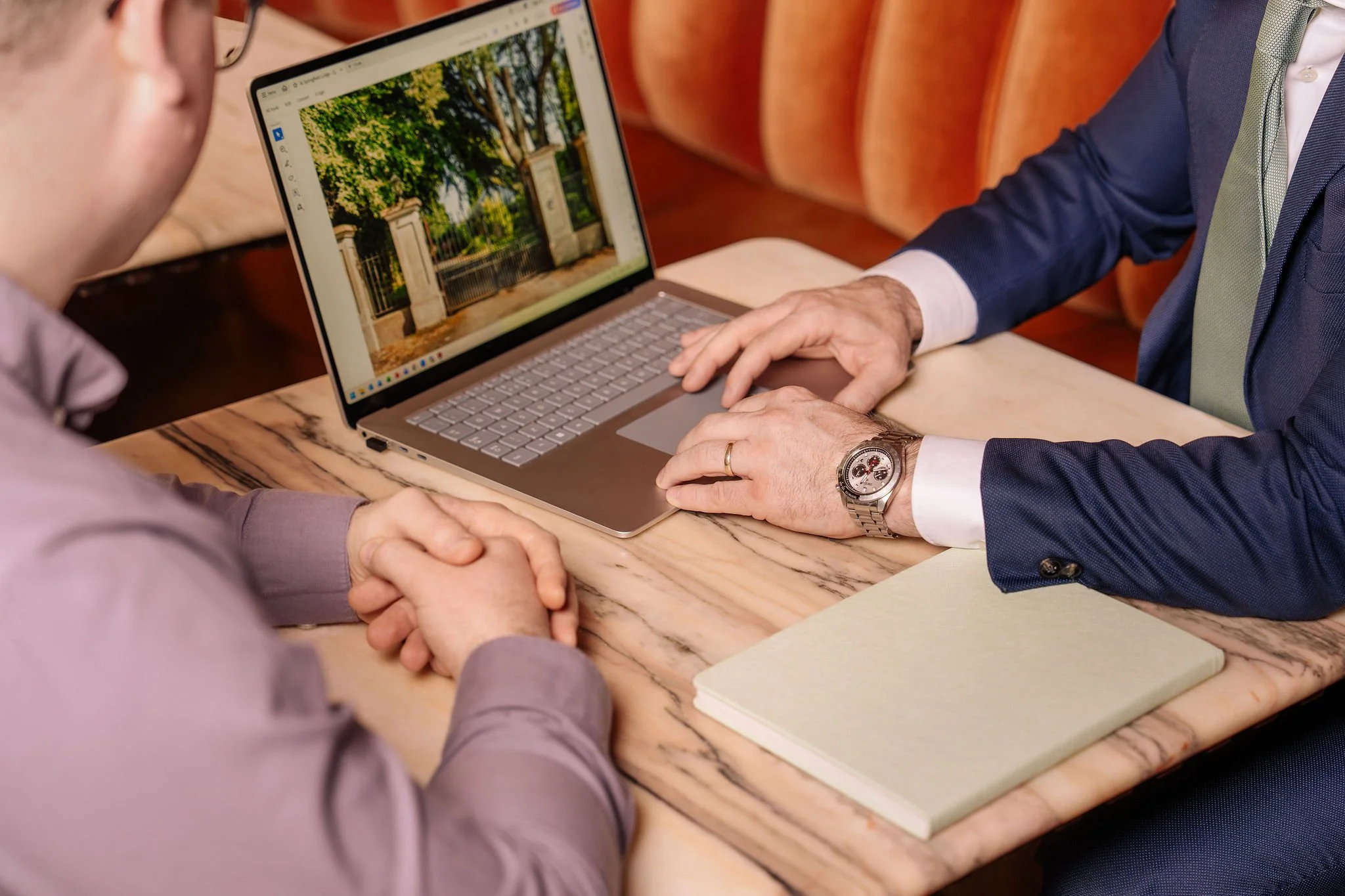 Two men are sitting at a wooden table having a discussion, with one man showing a picture of a park with trees on his laptop screen. One man is dressed in a suit with a watch and a wedding ring, while the other is wearing glasses and a light purple shirt. They are holding hands on the table.
