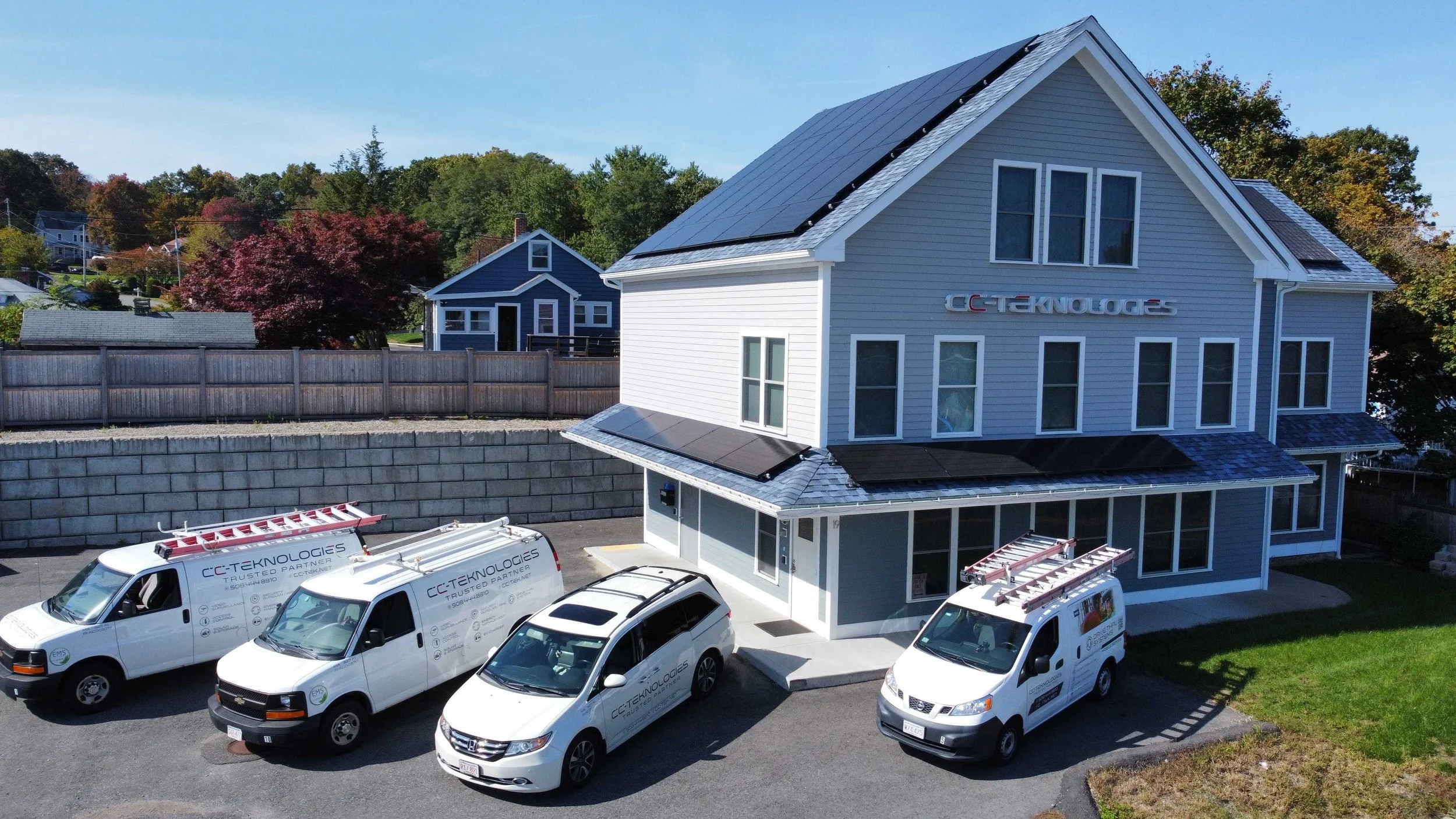 A light blue building with solar panels on the roof and a sign that reads 'CCTEKNOLOGIES'. Three white service vans and a white SUV are parked in front, all branded with 'CCTEKNOLOGIES' and related graphics.