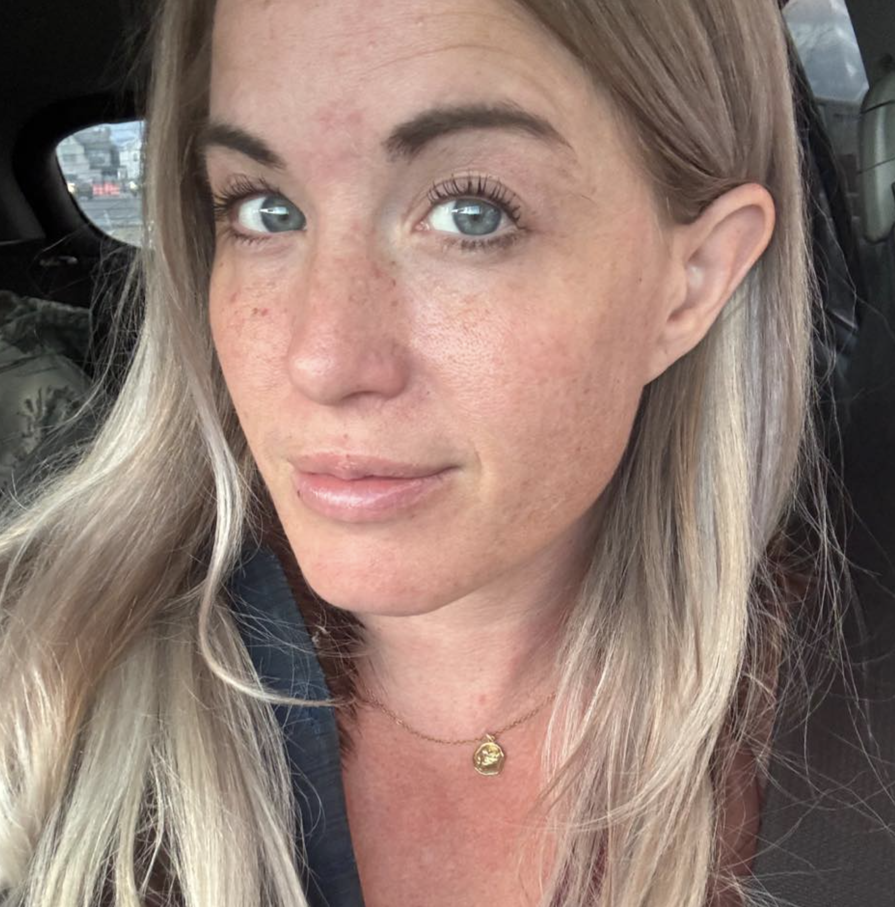 Nærbilde av en kvinne med langt blondt hår, blå øyne og smil. Hun har brukket øyenbrynene og noen fregner på ansiktet. Hun bærer en gull halskjede med en sirkelvedheng.