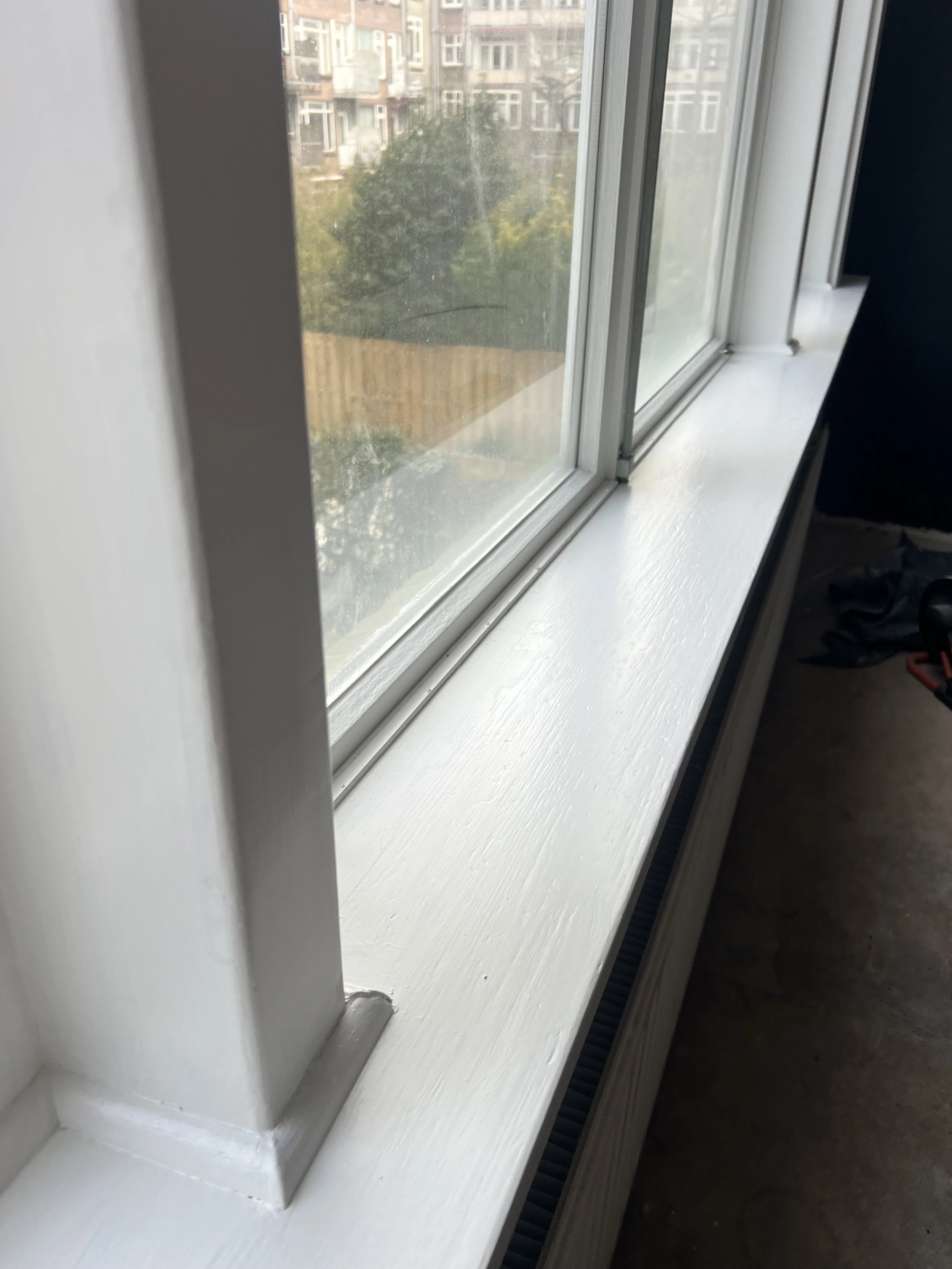 Nieuwer geschilderde witte vensterbank onder een raam met uitzicht op een tuin en gebouwen op de achtergrond.